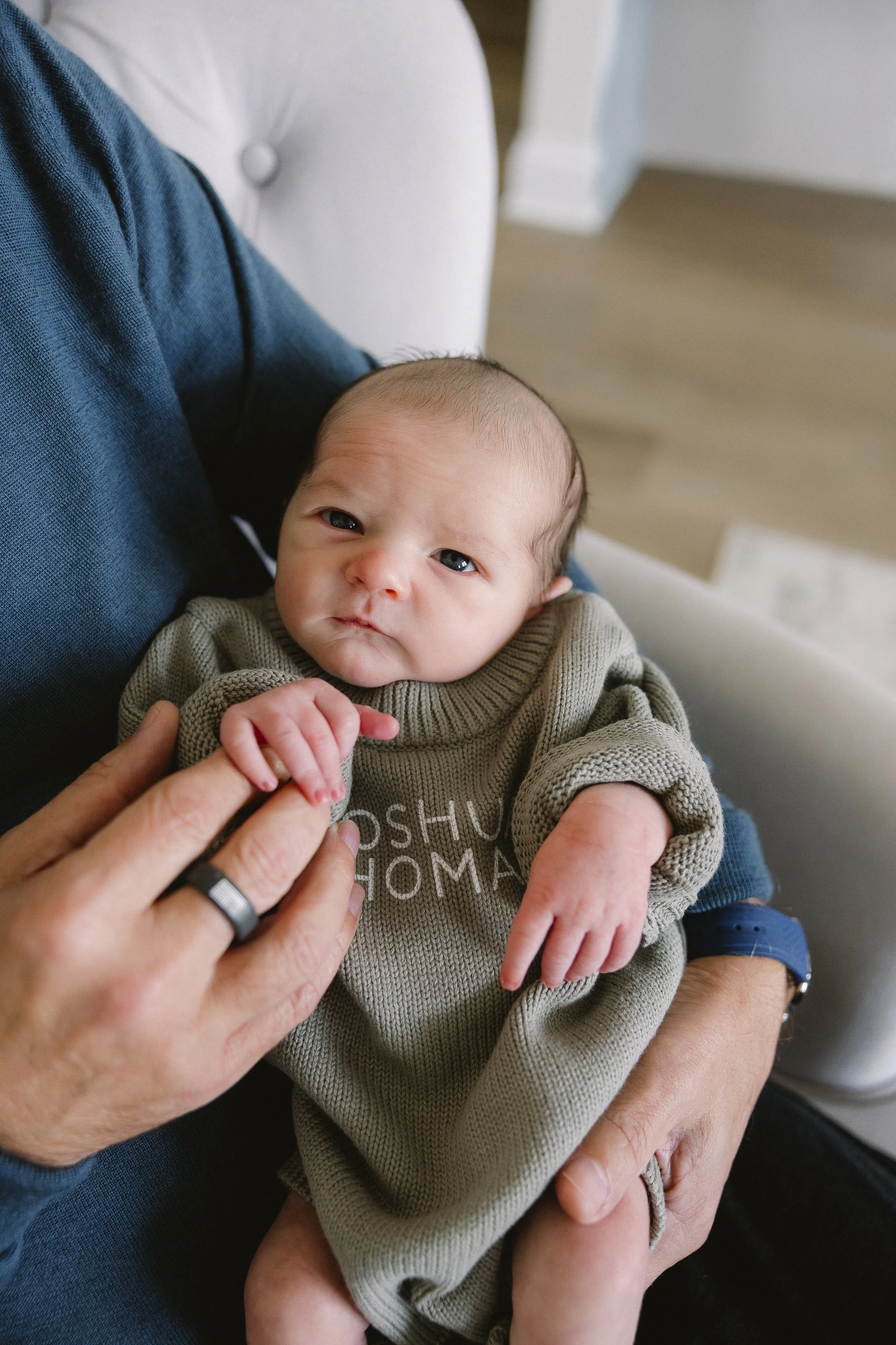 A baby with light skin and dark hair lying on an adult's lap, wearing a tan sweater with partially obscured text, and looking at the camera with a calm expression. Family Heirloom Photography with Storm Pierre Photography in Jacksonville NC.