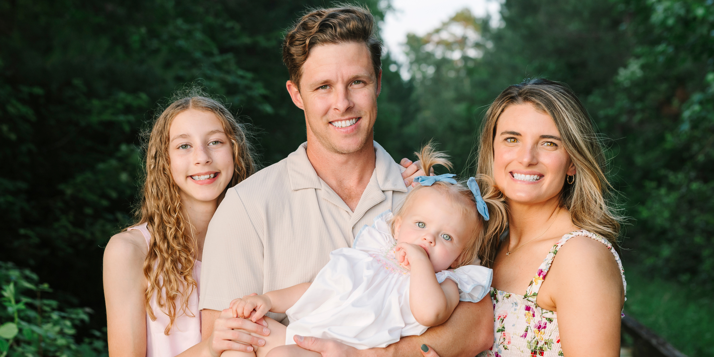 An outdoor family portrait featuring a father, mother, older daughter, and a baby girl posing closely together against a lush green forest background.