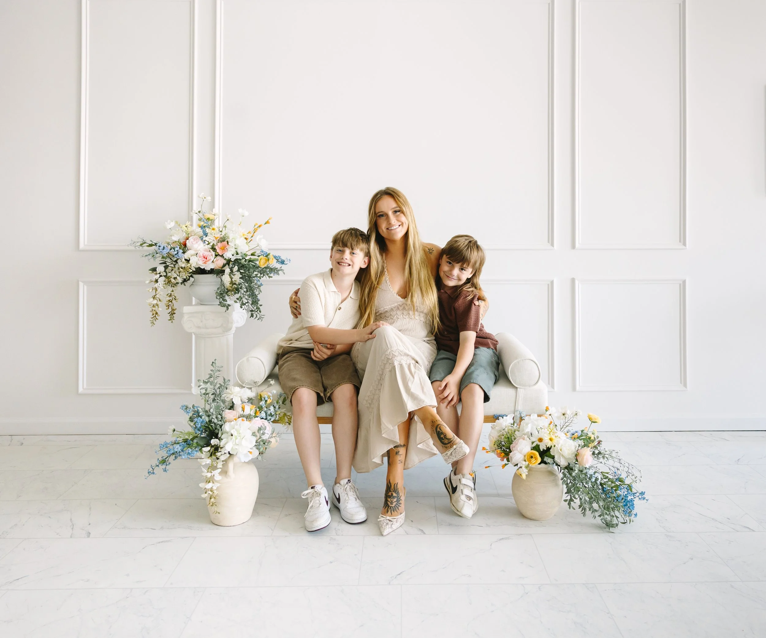 A woman with long blonde hair and tattoos on her legs sitting on a white sofa with two young boys, all smiling, Family Heirloom Photography with Storm Pierre Photography in Jacksonville NC serving Richlands, Swansboro, Hampstead, Sneads Ferry, Hubert