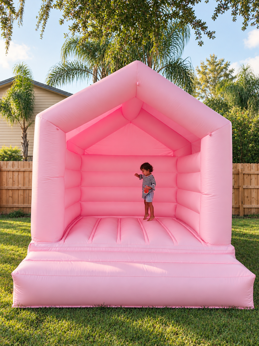 A young girl in a striped shirt and denim shorts playing inside a pink inflatable bounce house in a backyard with green grass, wooden fence, trees, and a house in the background.