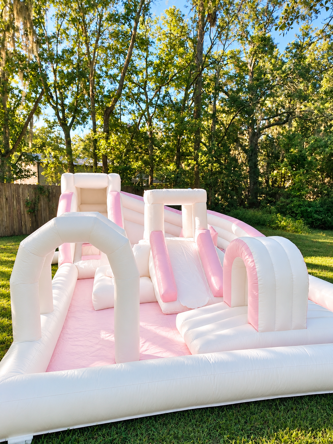 Pink and white inflatable bounce house with slides and climbing areas set up outdoors on a grassy lawn, surrounded by green trees and a wooden fence.