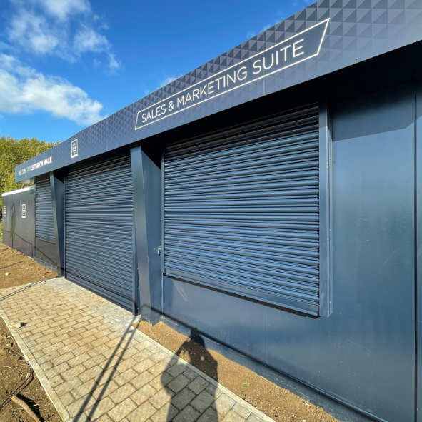 Exterior view of a building with closed rolling shutters and a signage that reads 'Sales & Marketing Suite' on a blue sky day.