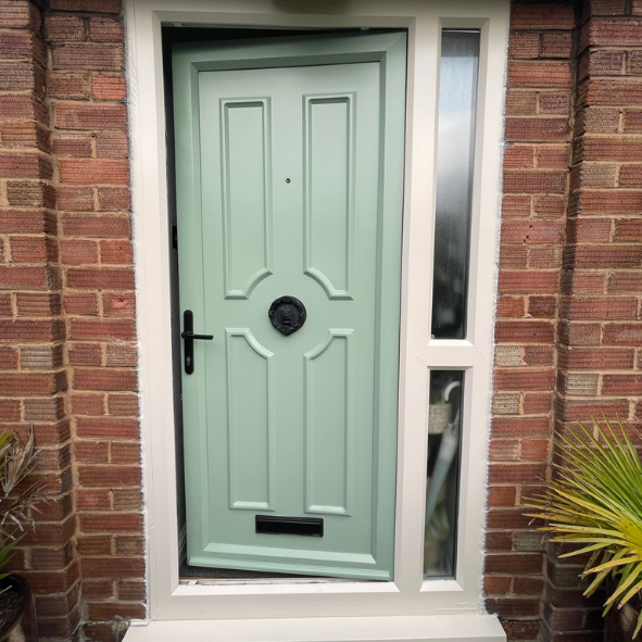 Front door with light green color, black door handle and mail slot, surrounded by red brick wall and narrow window on the side, with a potted plant on the right.