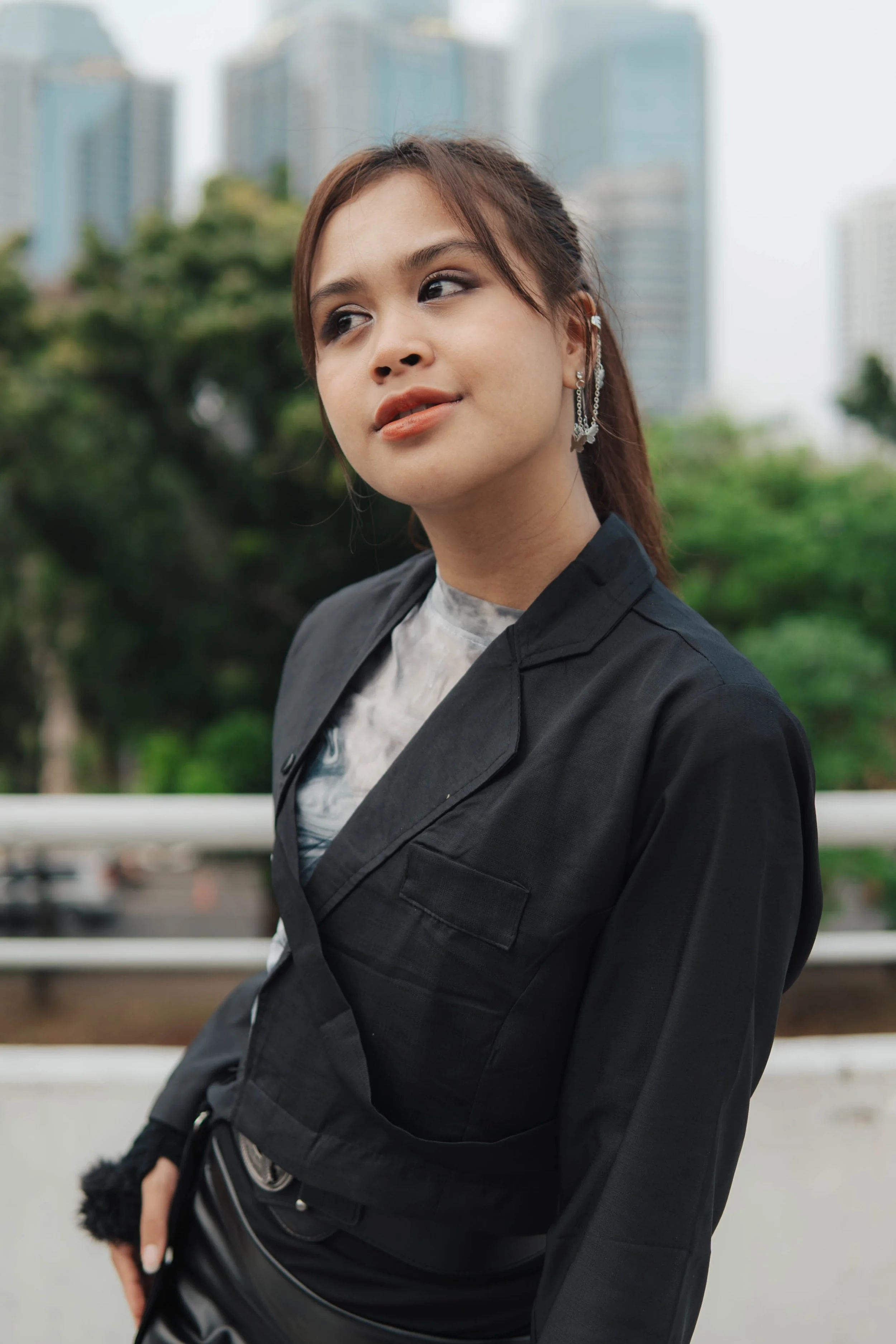 A woman wearing a black jacket and earrings, standing outdoors with city buildings and trees in the background.