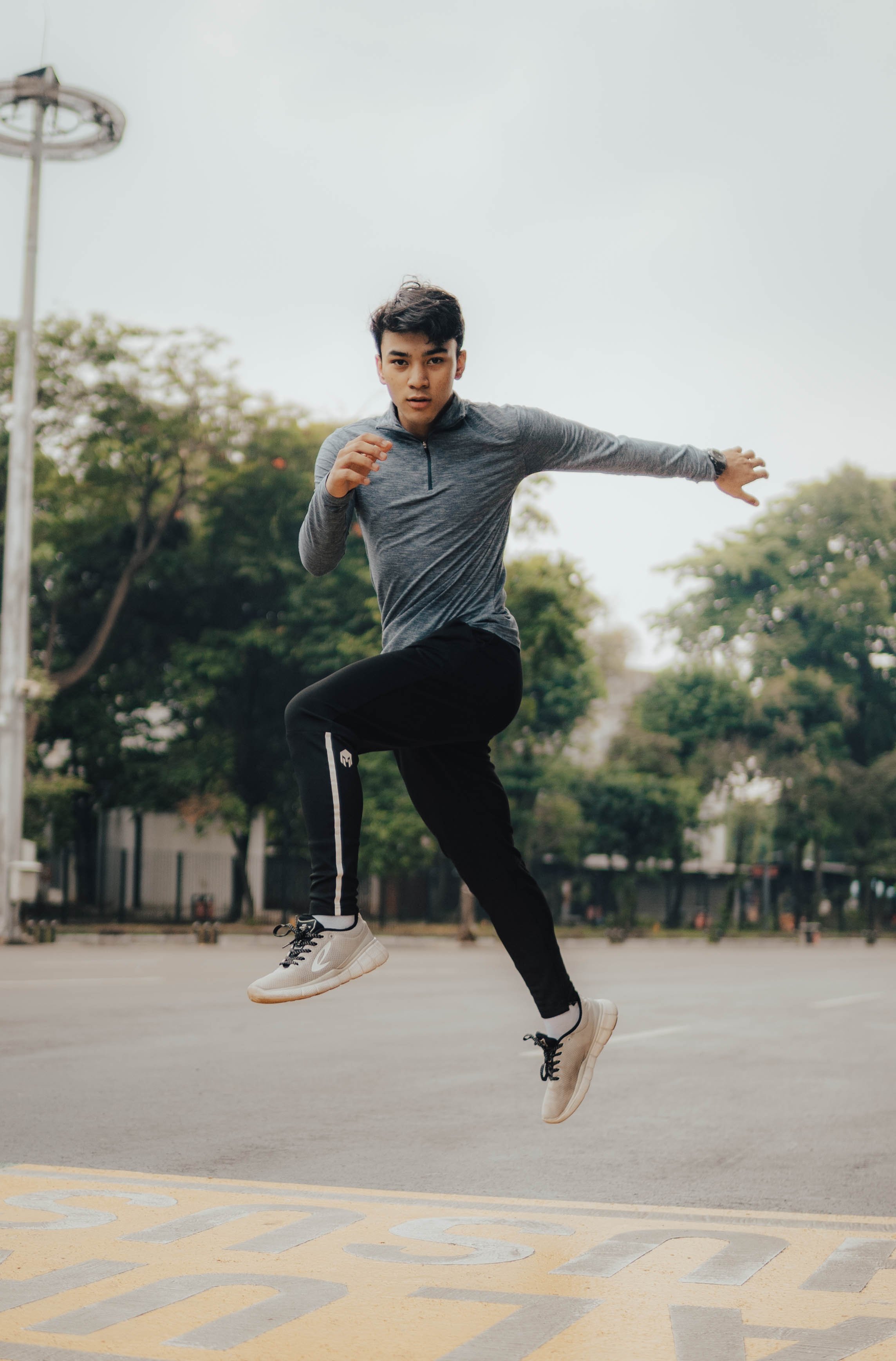 Man in athletic clothing jumping outdoors on a street with trees in the background.