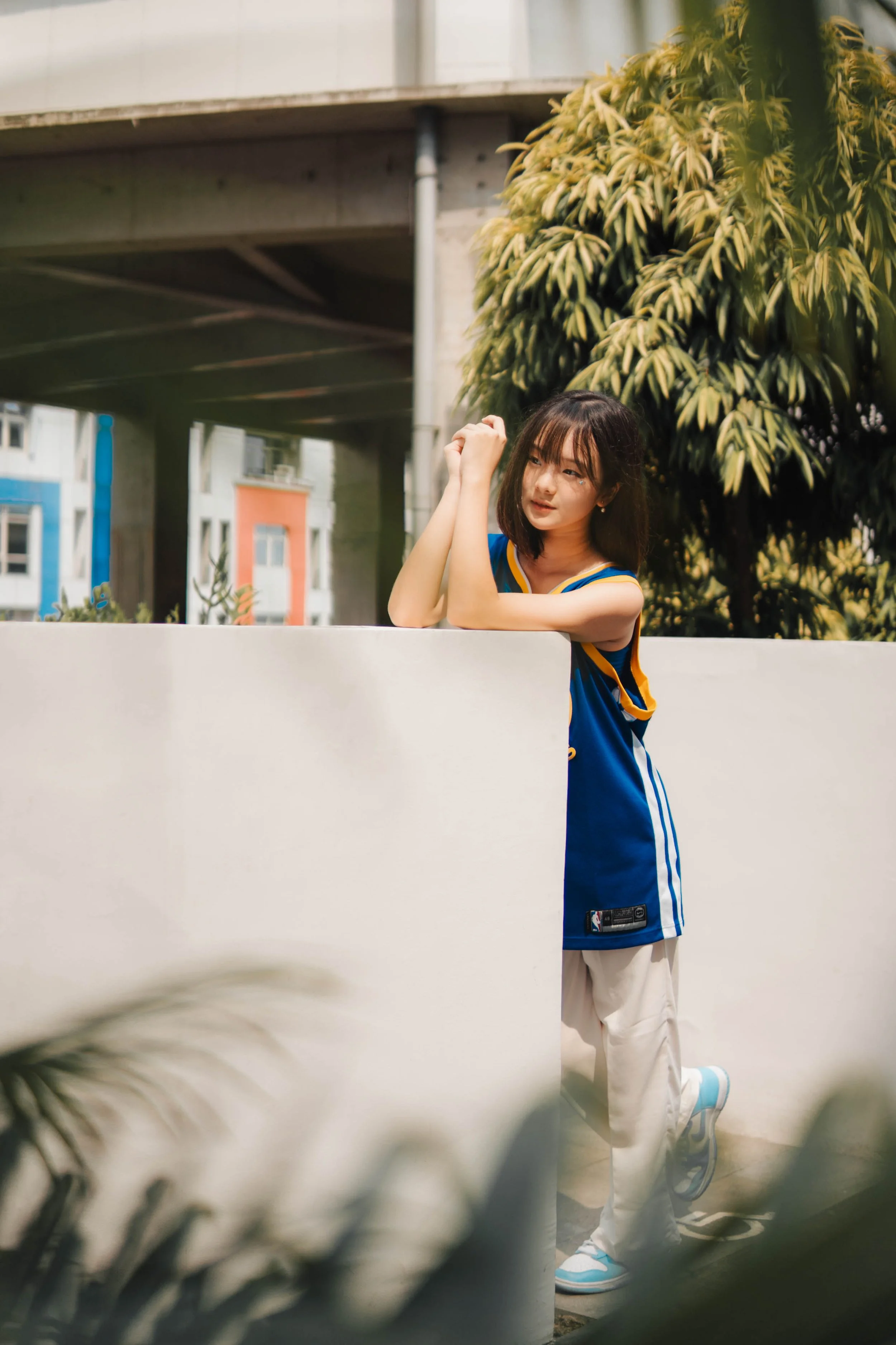 A person wearing a blue sports jersey and white pants, leaning on a white wall outdoors, with trees and buildings in the background.