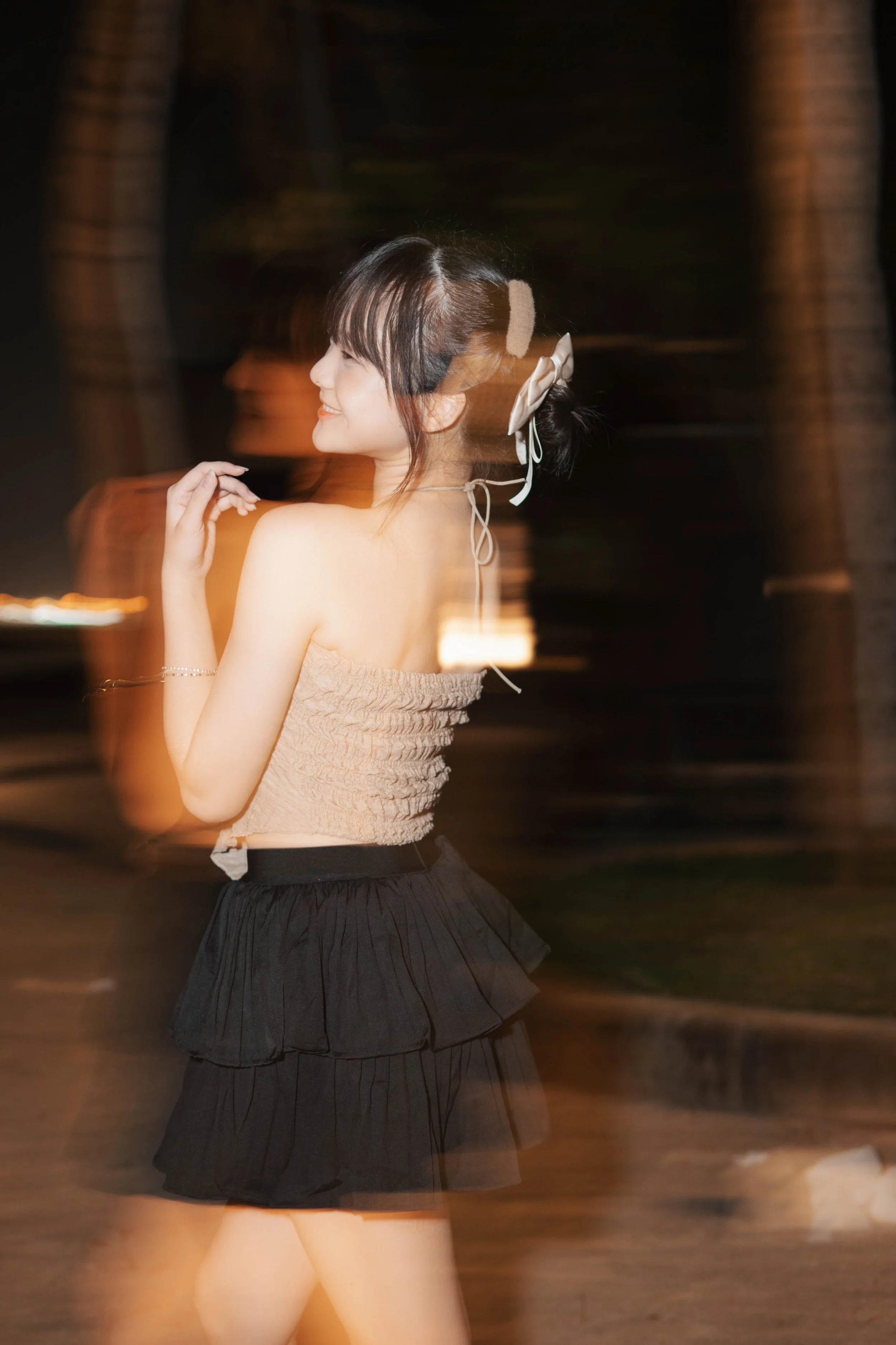 A woman in a beige tube top and black skirt, outdoors at night, slightly blurred motion effect.