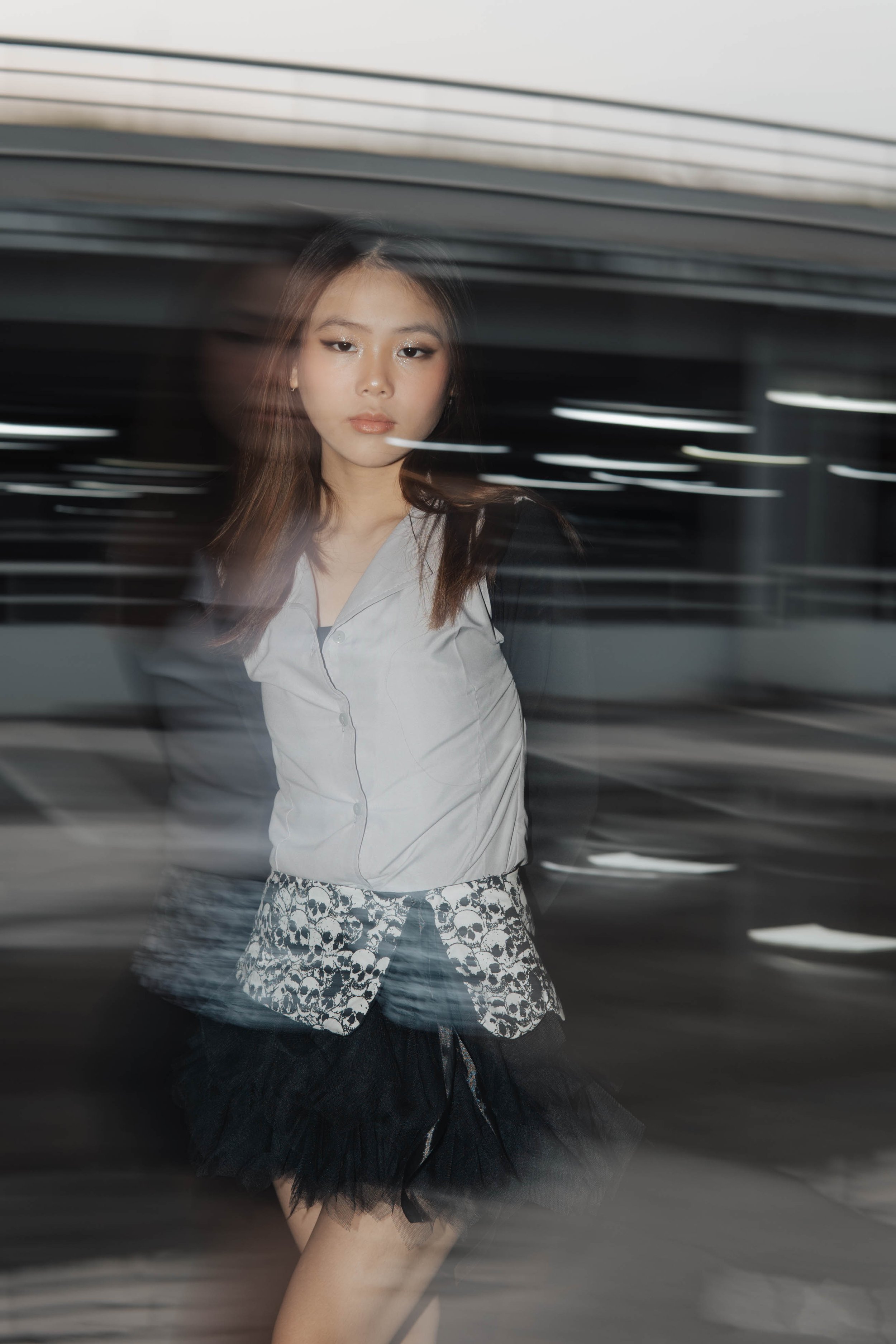 Blurred image of a person standing in an urban setting with motion blur effect, wearing a light-colored shirt and a skirt with skull patterns.