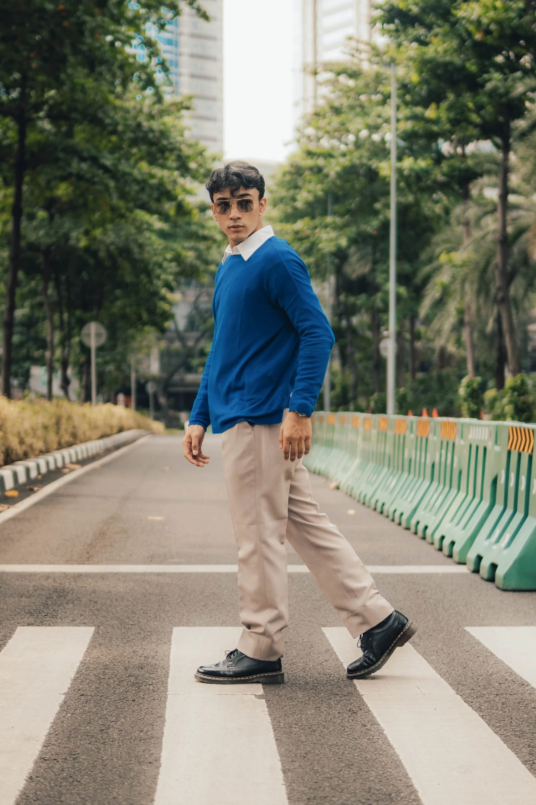 A person wearing sunglasses, a blue sweater, and beige pants walks on a crosswalk in an urban area with trees and barriers in the background.