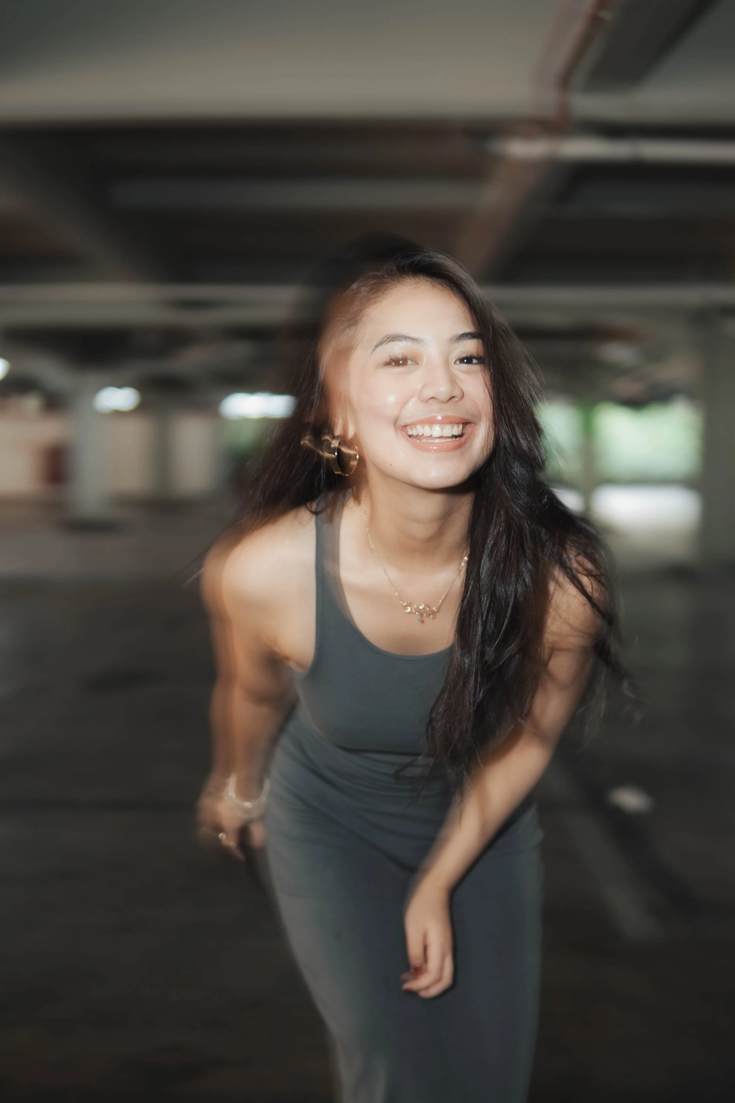 A smiling woman leaning forward in a dimly lit indoor space, wearing a dark sleeveless dress with long hair.