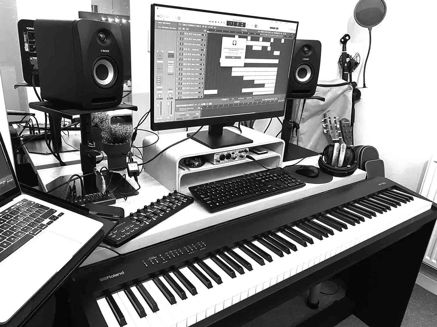 Anne's home recording studio with a digital audio workstation, a monitor, studio monitor speakers, midi cc controller, a digital keyboard, a laptop, headphones, and other audio equipment on a desk.