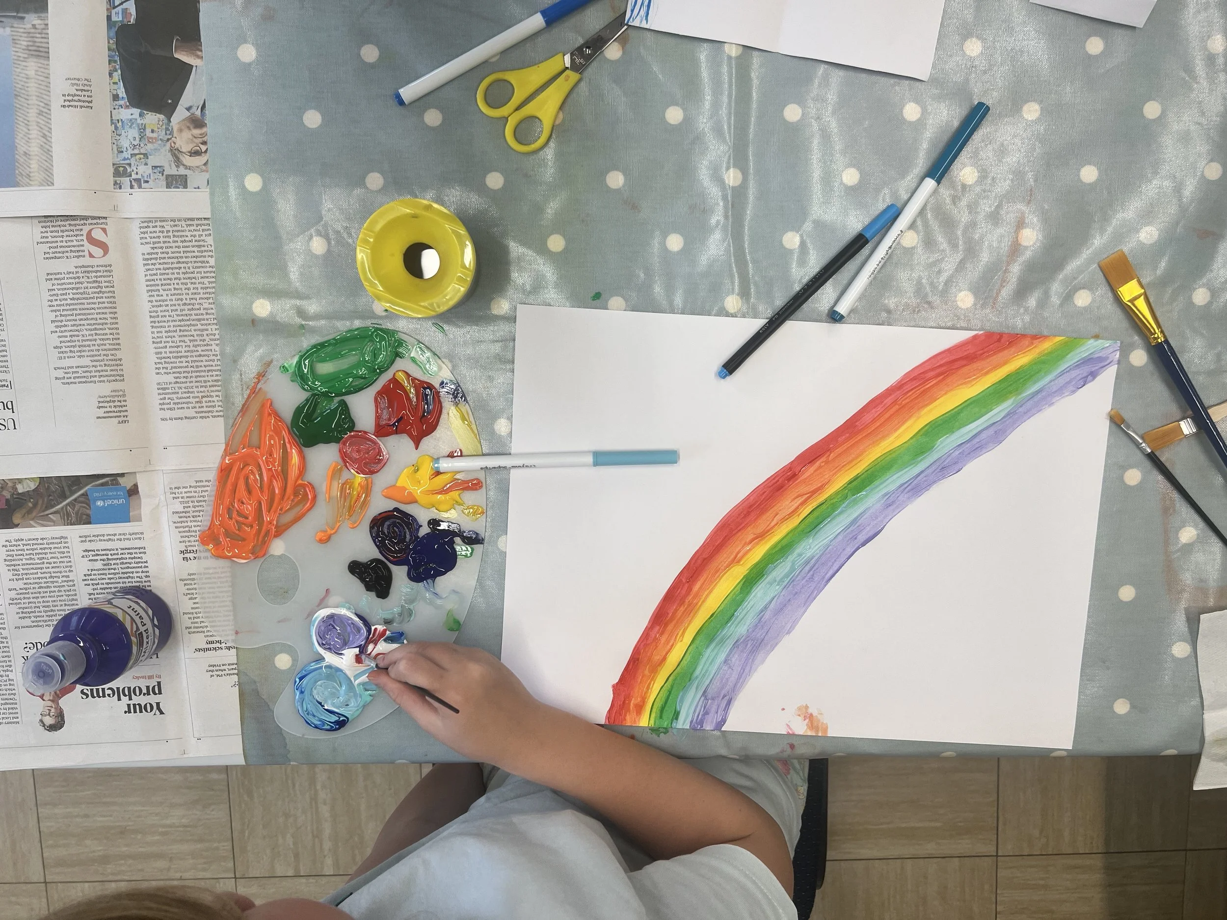 A child's hand painting a rainbow on a large white sheet of paper. The rainbow is painted in the colors red, orange, yellow, green, blue, and purple. The table has various art supplies, including paint, brushes, a palette, markers, a glue stick, and scissors. Newspapers are spread on the table to protect it from paint.