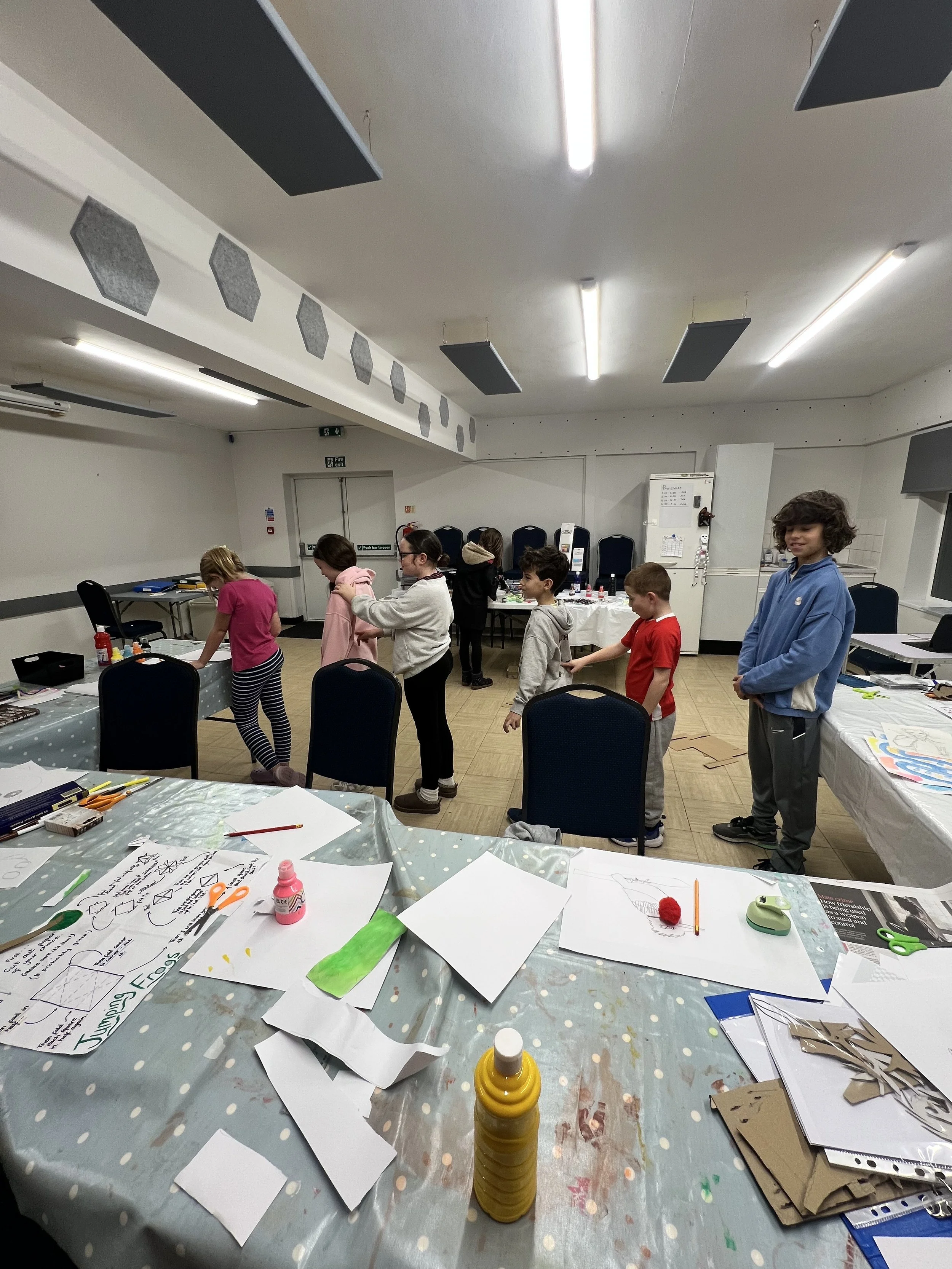 Children participating in an indoor arts and crafts activity in a classroom, with tables covered in paper, art supplies, and completed projects.