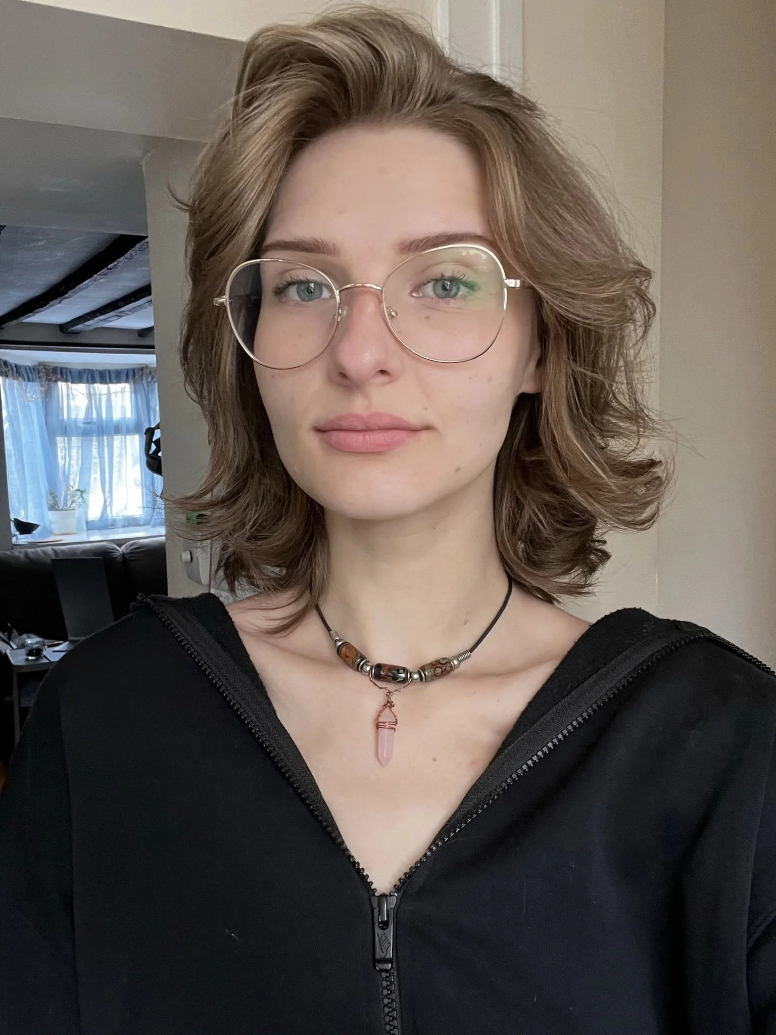 A young woman with shoulder-length wavy brown hair wearing glasses, a black zip-up hoodie, and a beaded necklace with a pink crystal pendant, standing indoors in front of a beige wall.
