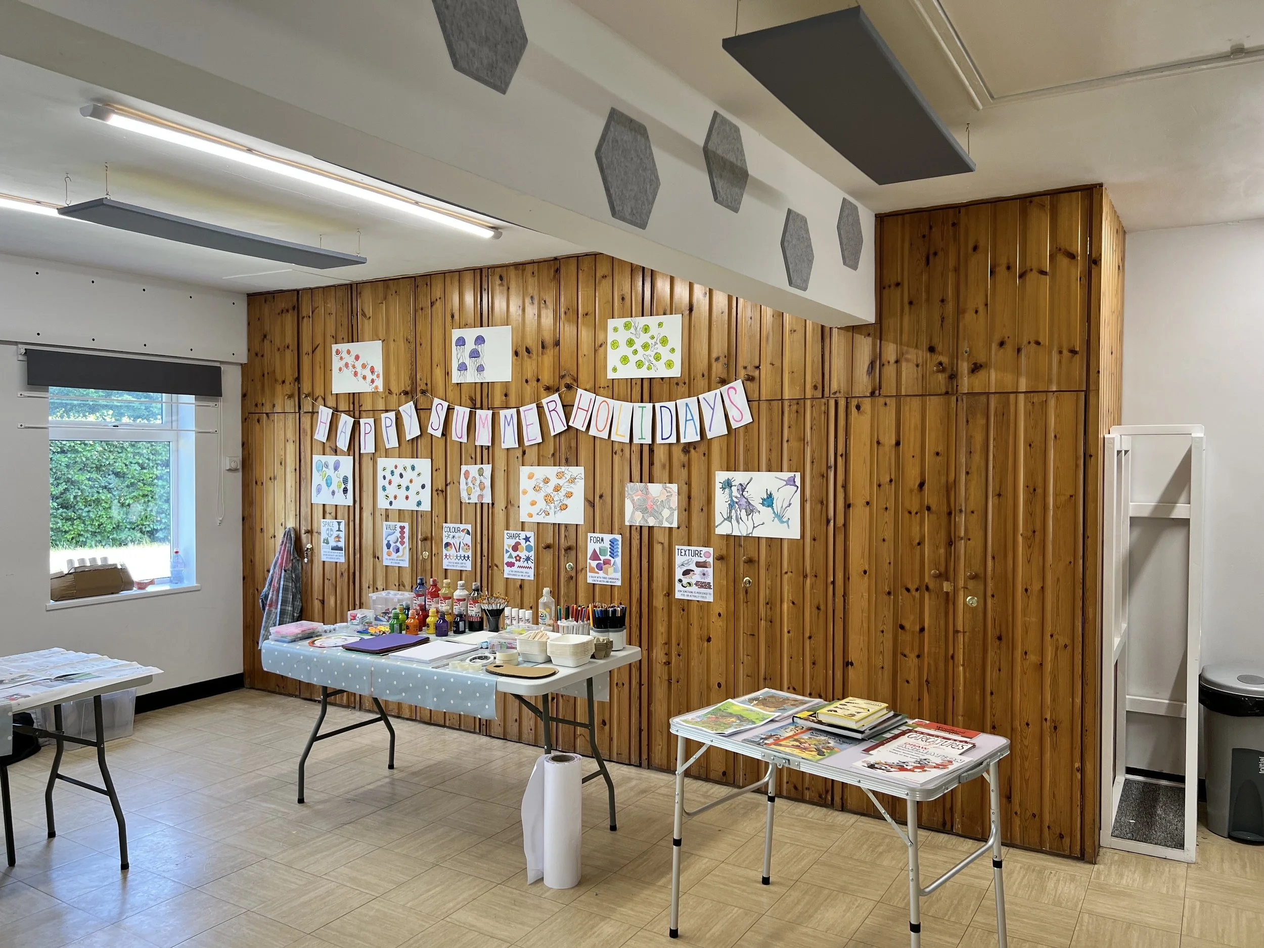 A decorated classroom with a wooden wall displaying colorful artwork and a banner that reads 'Happy Summer Holidays.' Tables hold art supplies and magazines, and there is a window with a view of greenery outside.