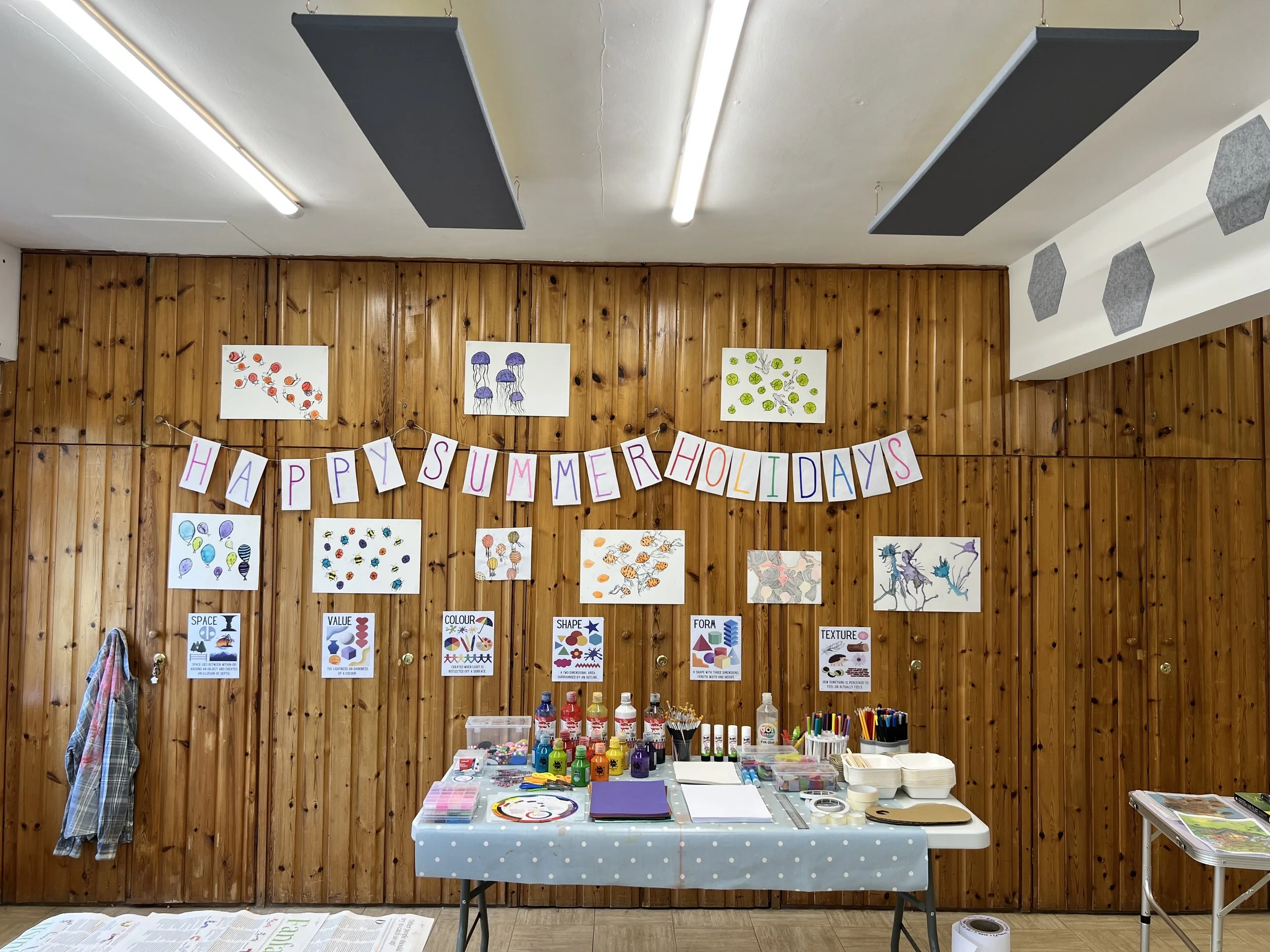 A decorated room with a wooden wall and colorful artwork and banners. A table with art supplies is set up in front of the wall, and a banner reads "Happy Summer Holidays."