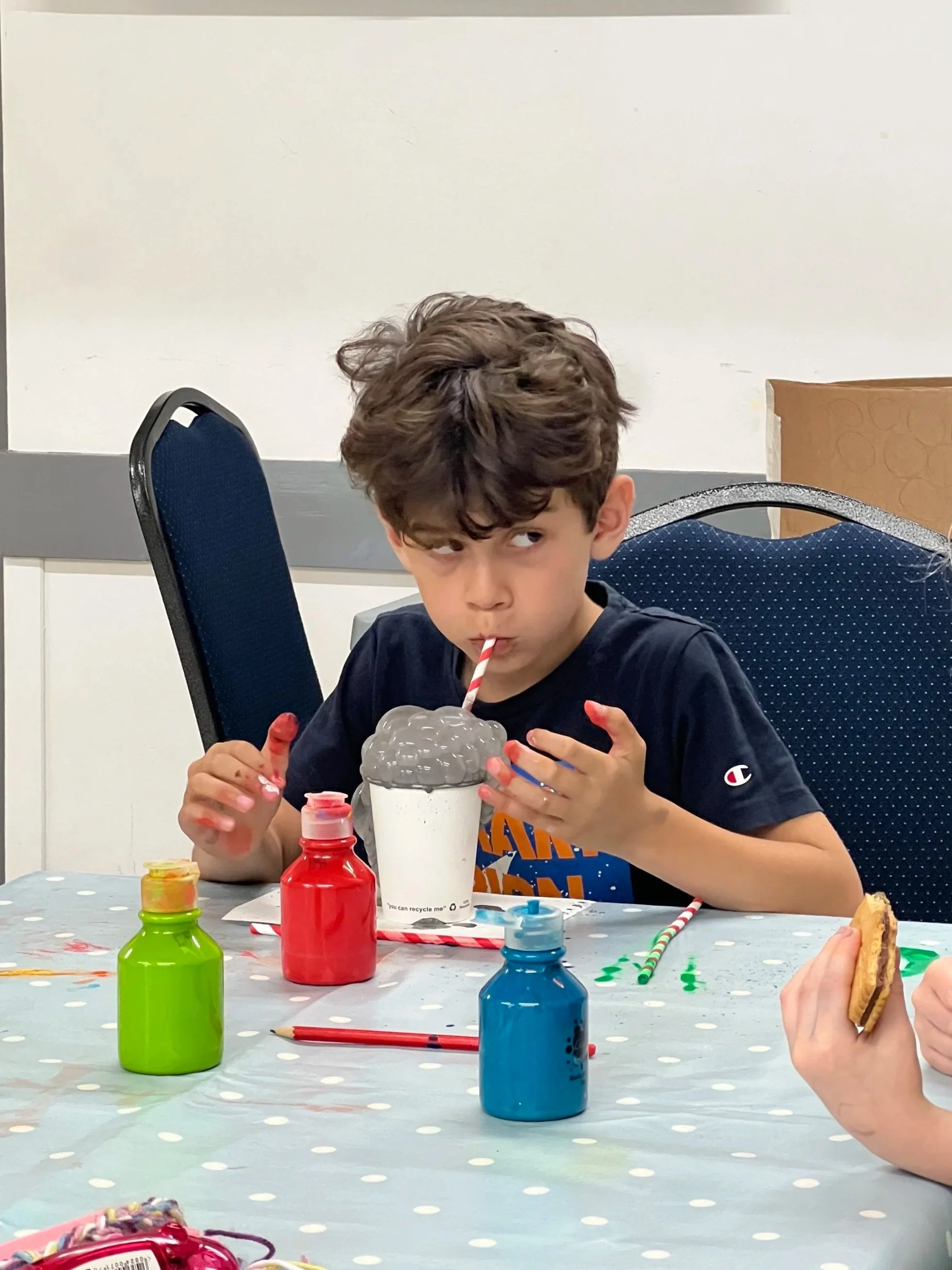 Young boy with messy hair drinking a cotton candy soda through a red and white striped straw, sitting at a table decorated with paint bottles and colored pencils, and holding a cookie in one hand.