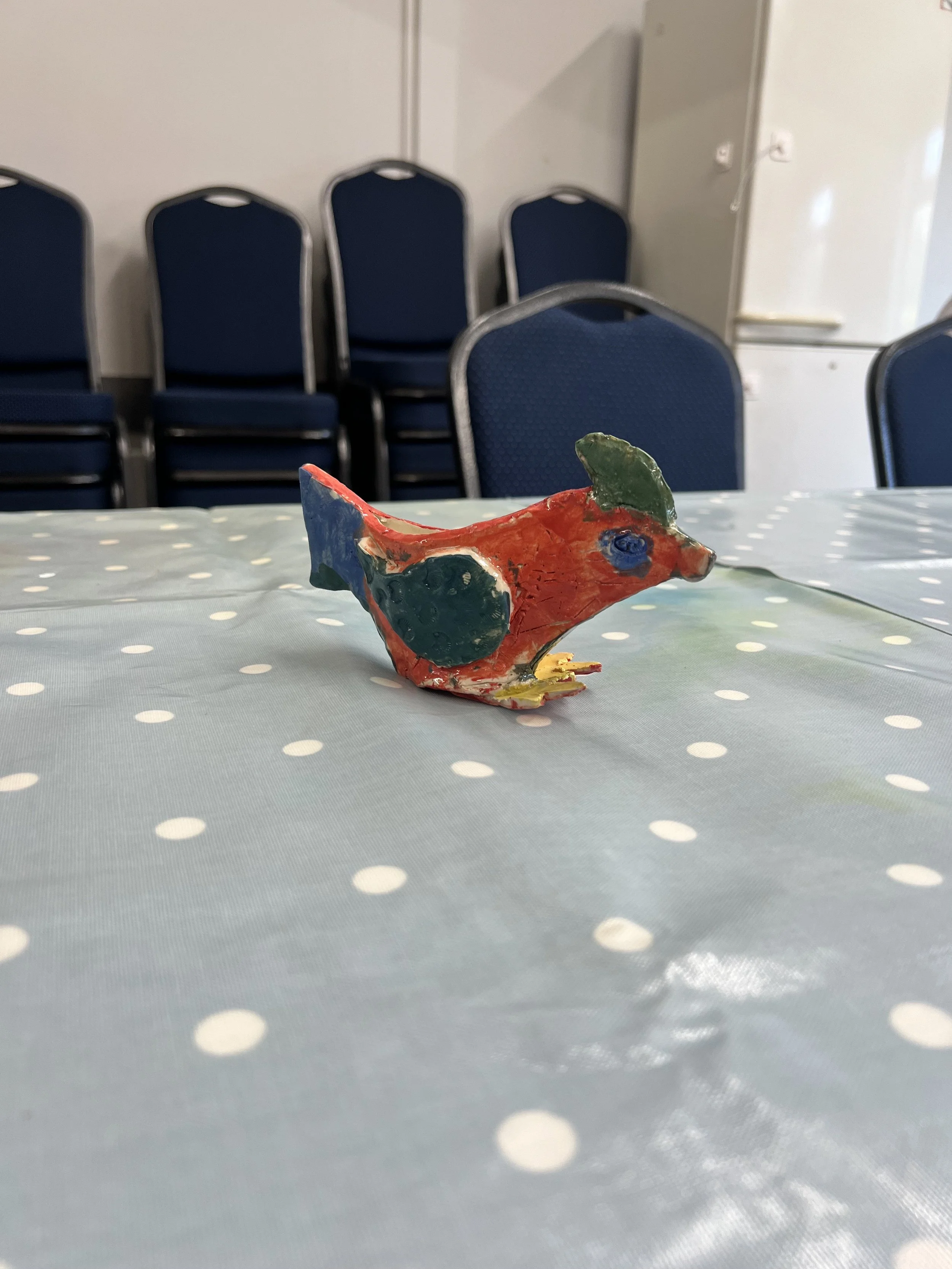 Colorful ceramic bird figurine on a polka dot tablecloth with stacked chairs and a cabinet in the background.