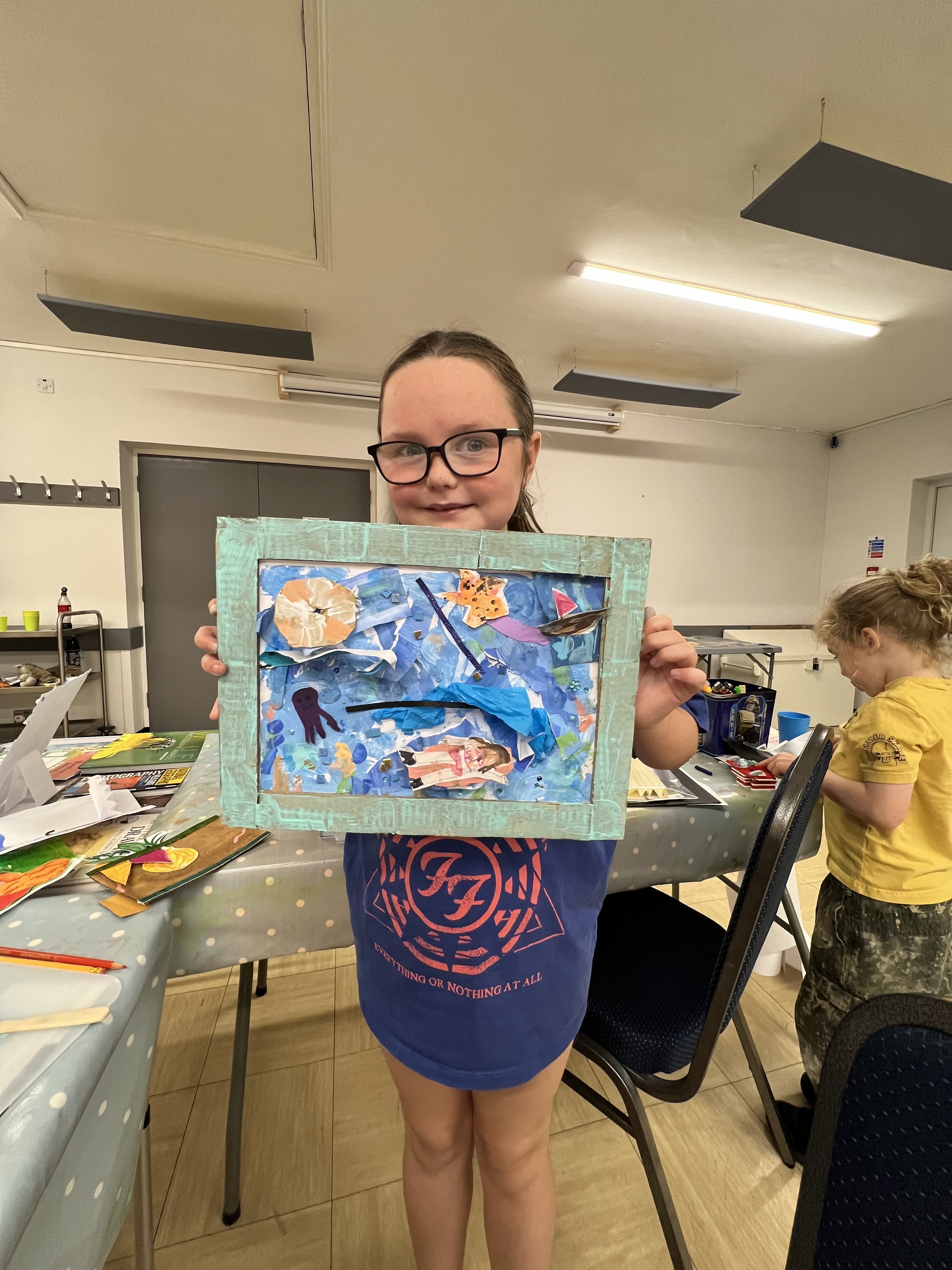 Young girl holding a homemade craft picture with ocean and sea-themed collages, at a table covered with art supplies in a classroom.