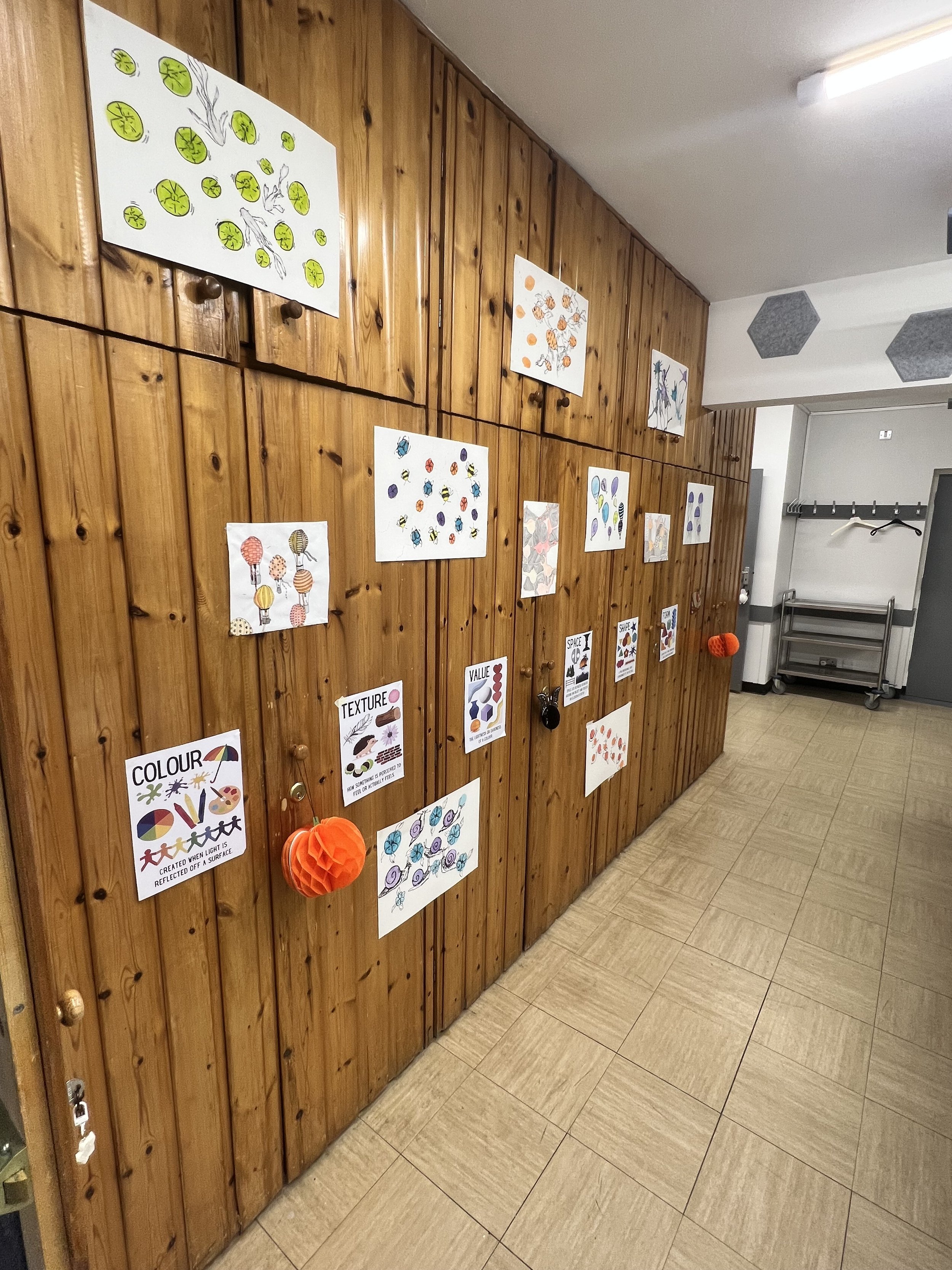 Wall of wooden lockers decorated with children's educational artwork and posters about colors, textures, and values, with orange paper decorations hanging on some lockers.