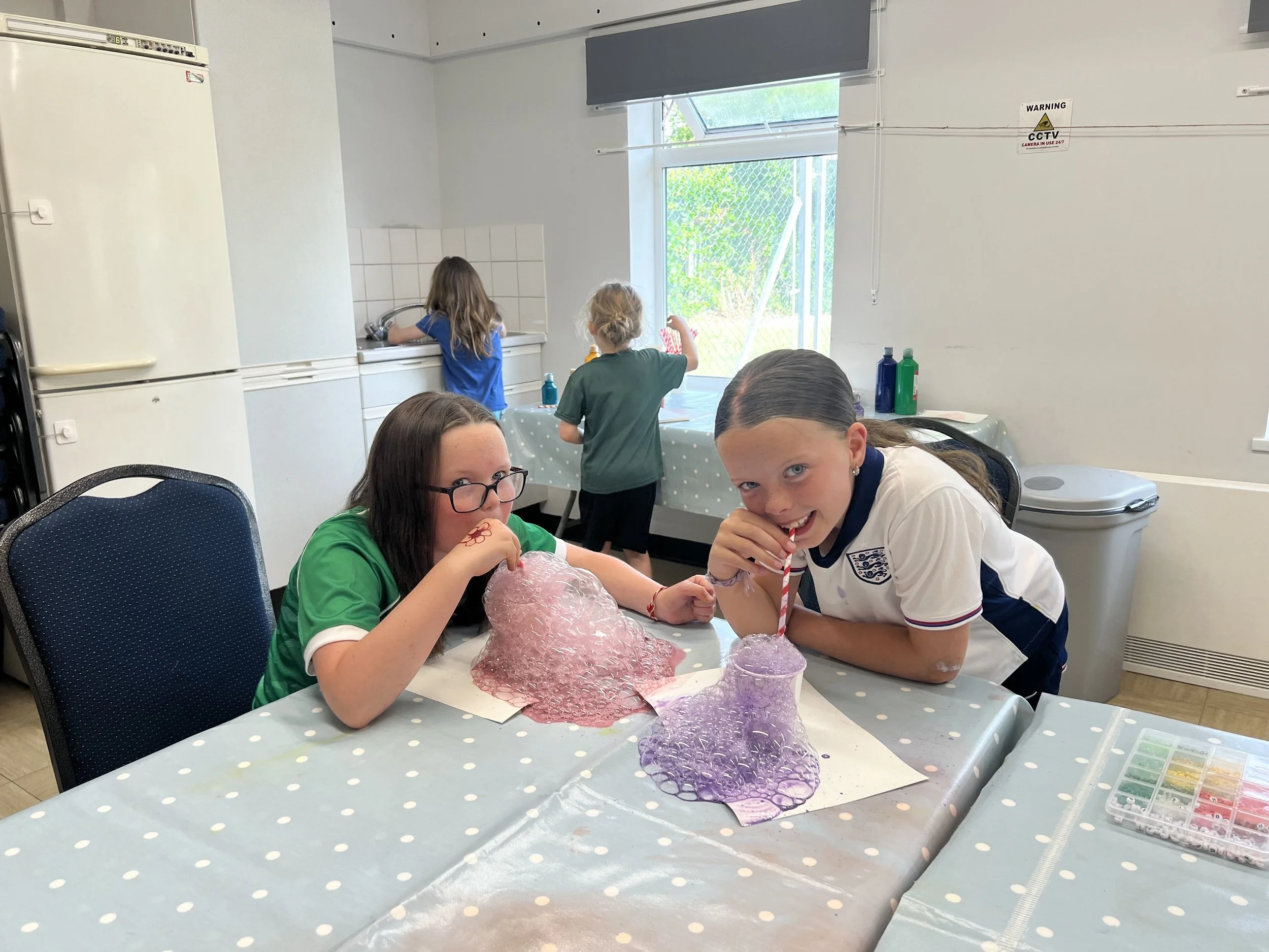 Two girls blowing bubbles at a table during a craft activity in a room, with three children in the background near the window and sink.