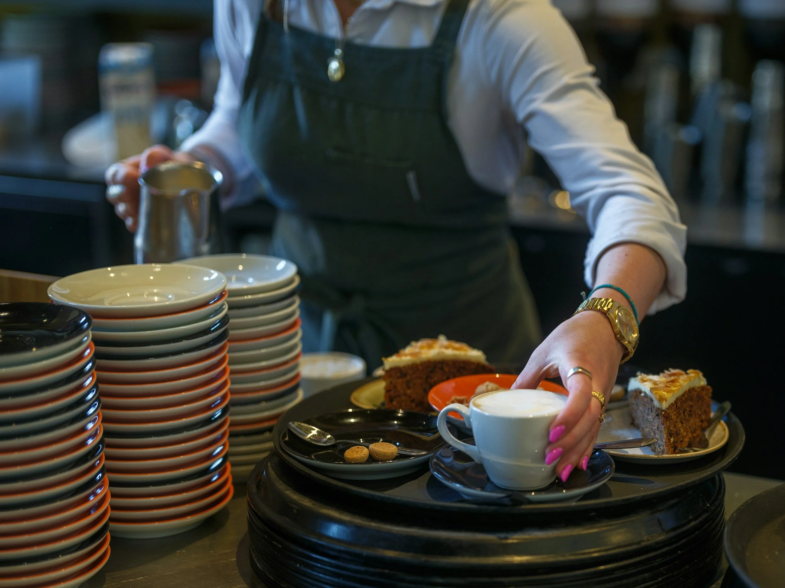 A person wearing a white shirt and dark apron is serving coffee and slices of cake in a cafe. There are stacked bowls and plates on the counter.