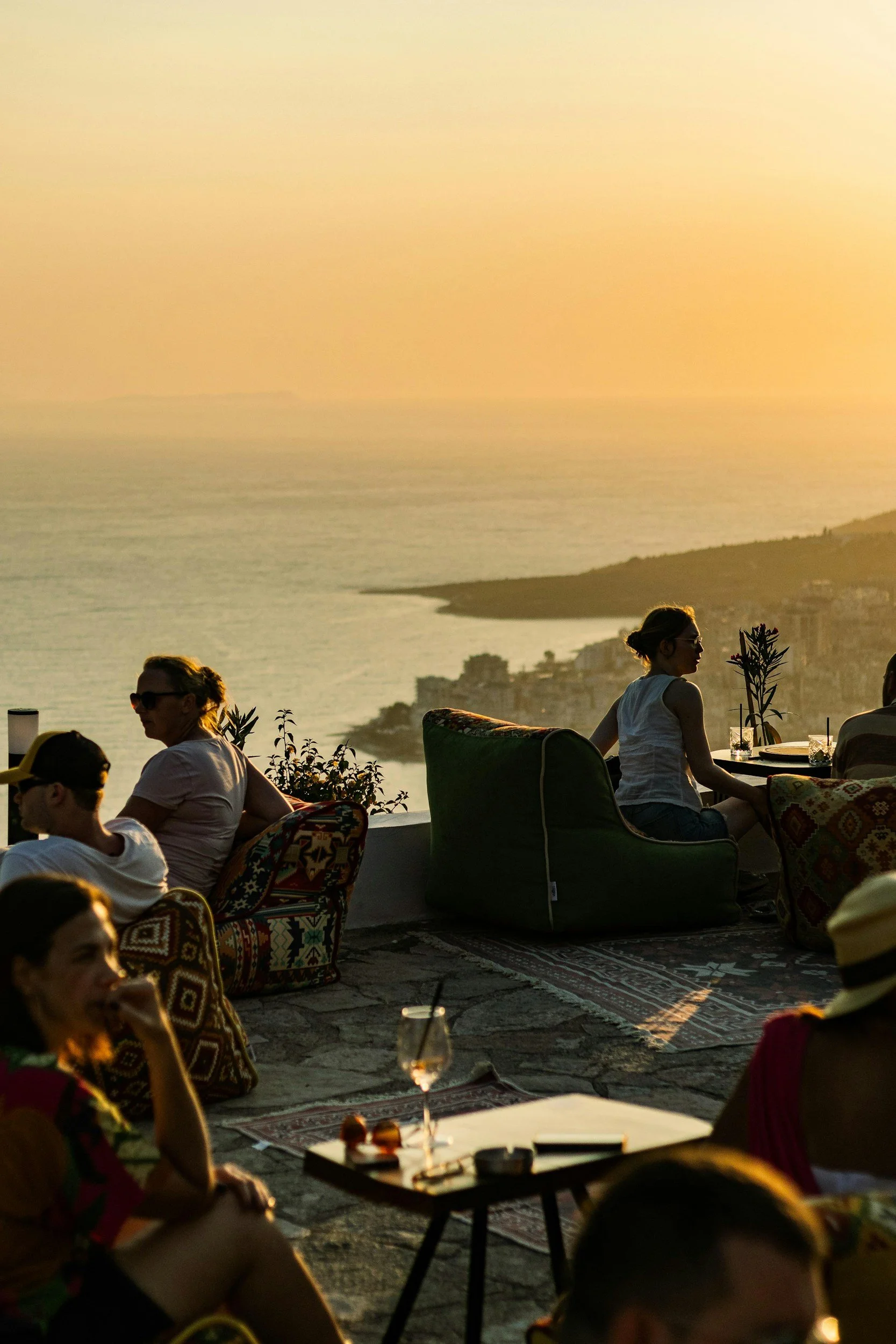 People sitting outdoors on a terrace overlooking a body of water during sunset, with some holding drinks and engaging in conversation.