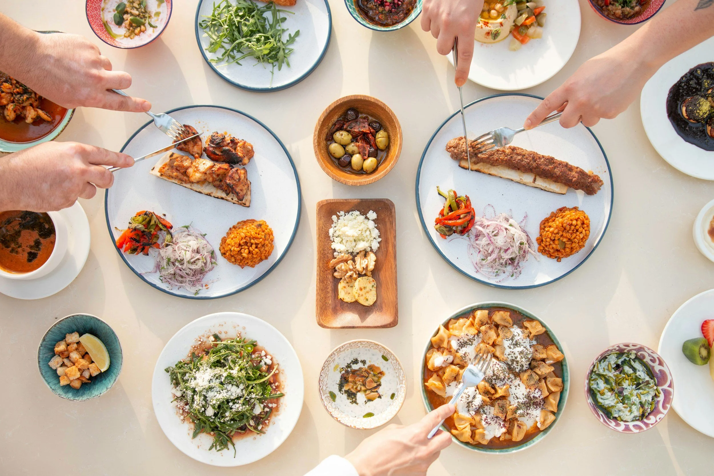 A top-down view of a table filled with various dishes of a meal, including slices of pizza, salads, sauces, and small bowls with a variety of food items. Several hands are visible, using forks and knives to serve themselves.
