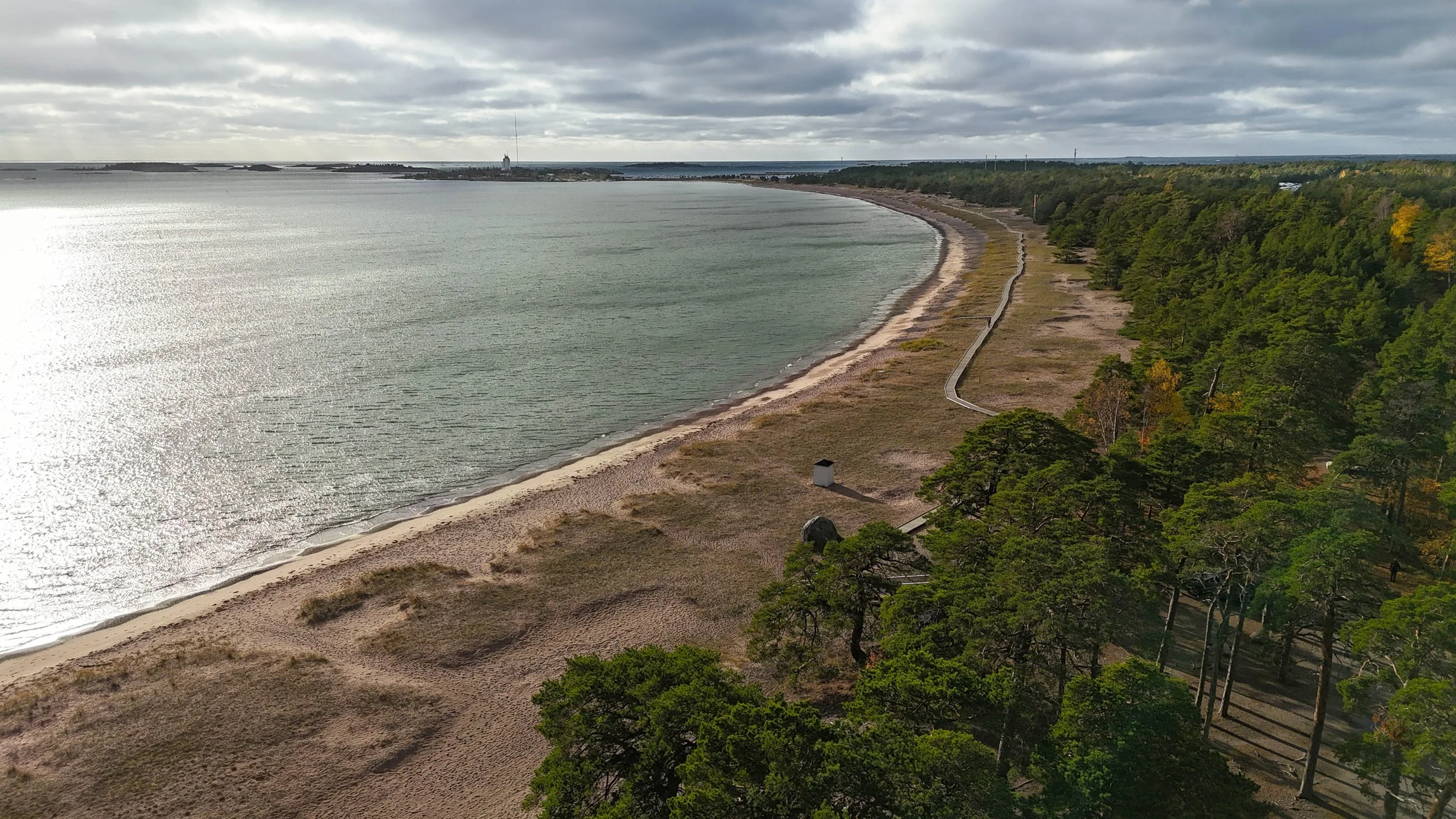 Aerial view of a long, curved beach with sandy shores, surrounded by dense green forests, with cloudy skies overhead and a body of water extending to the horizon.