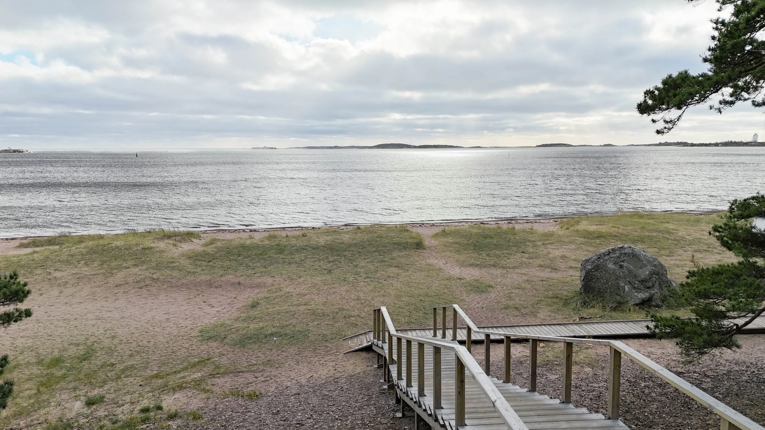 A leading down view of a beach with sand, grass, and a small wooden ramp leading to the shoreline, with calm water and a cloudy sky in the distance.
