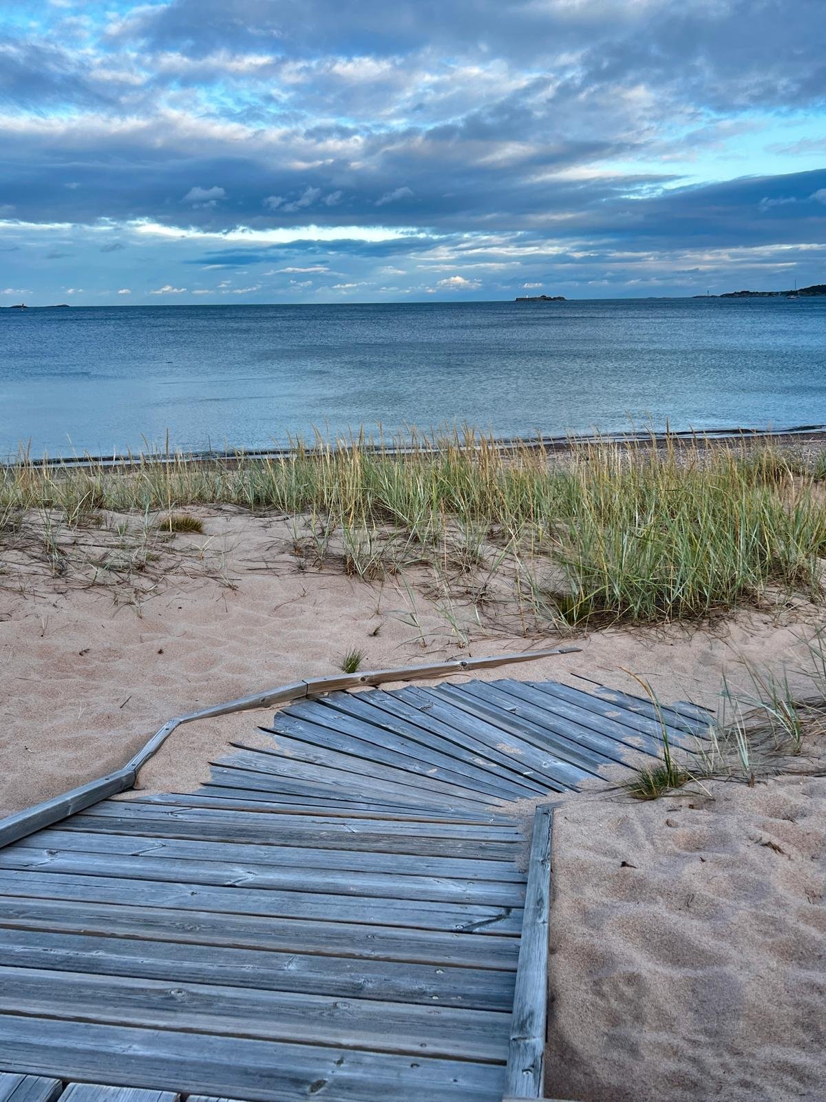 A sandy beach with a wooden boardwalk leading to the water, with tall grass along the shoreline. The ocean stretches out to the horizon with a cloudy sky overhead and a distant landmass.