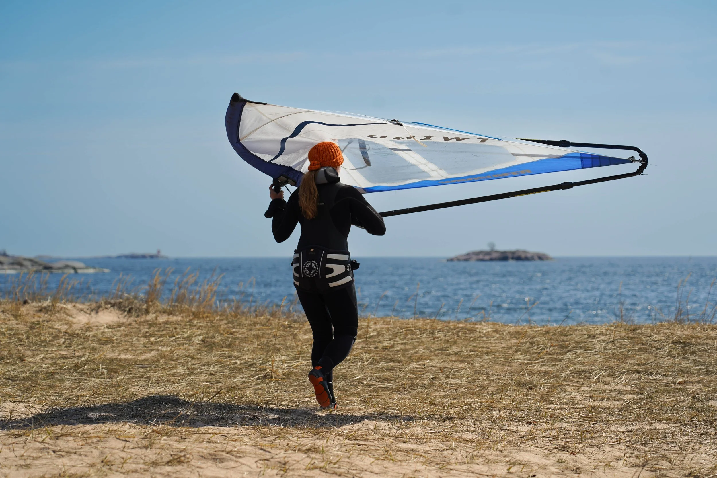 Person running on sandy beach carrying a large windsurfing sail with the ocean and small island in the background.