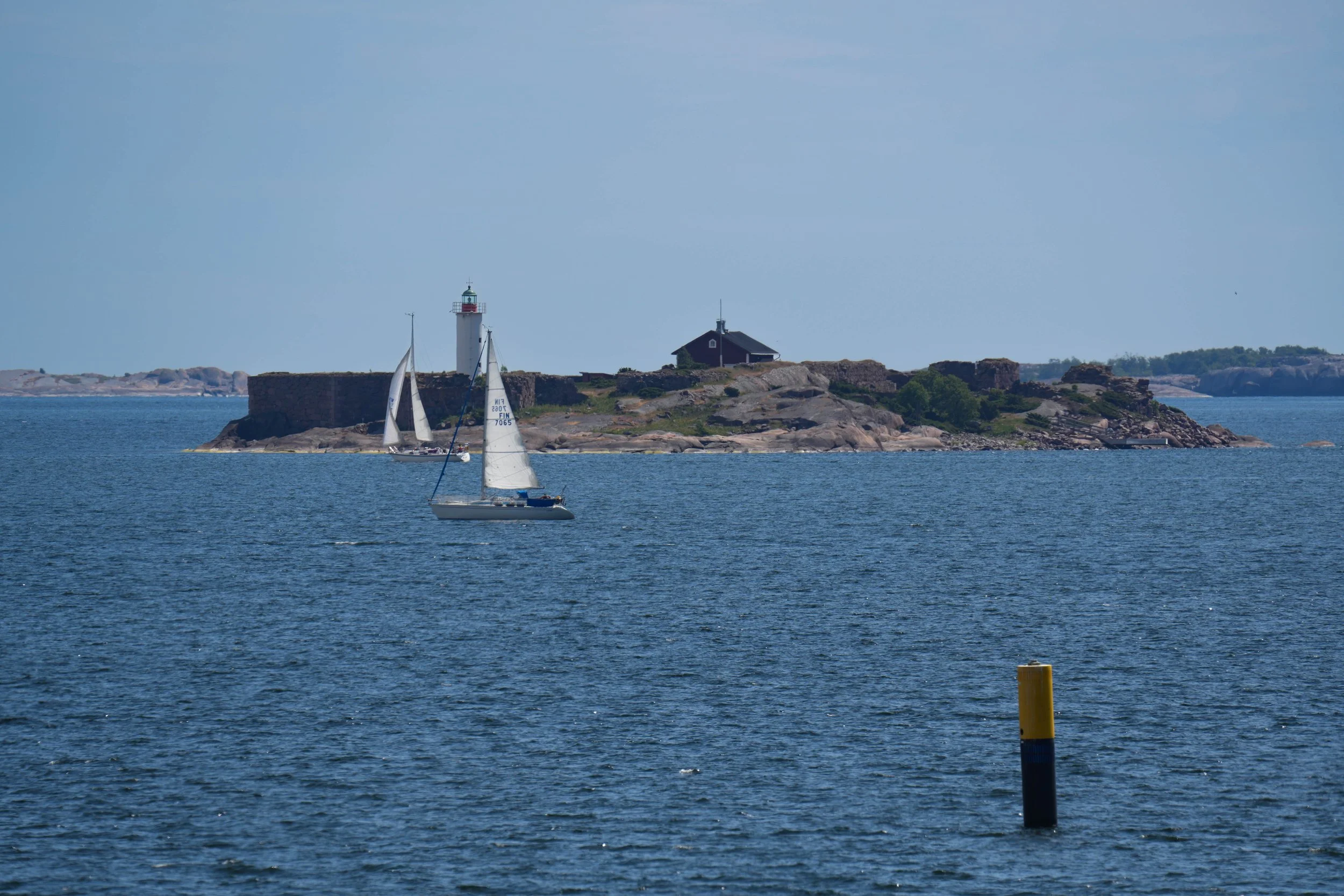 Two sailboats on the water near a rocky island with a lighthouse and a building, with a yellow and black buoy in the foreground.