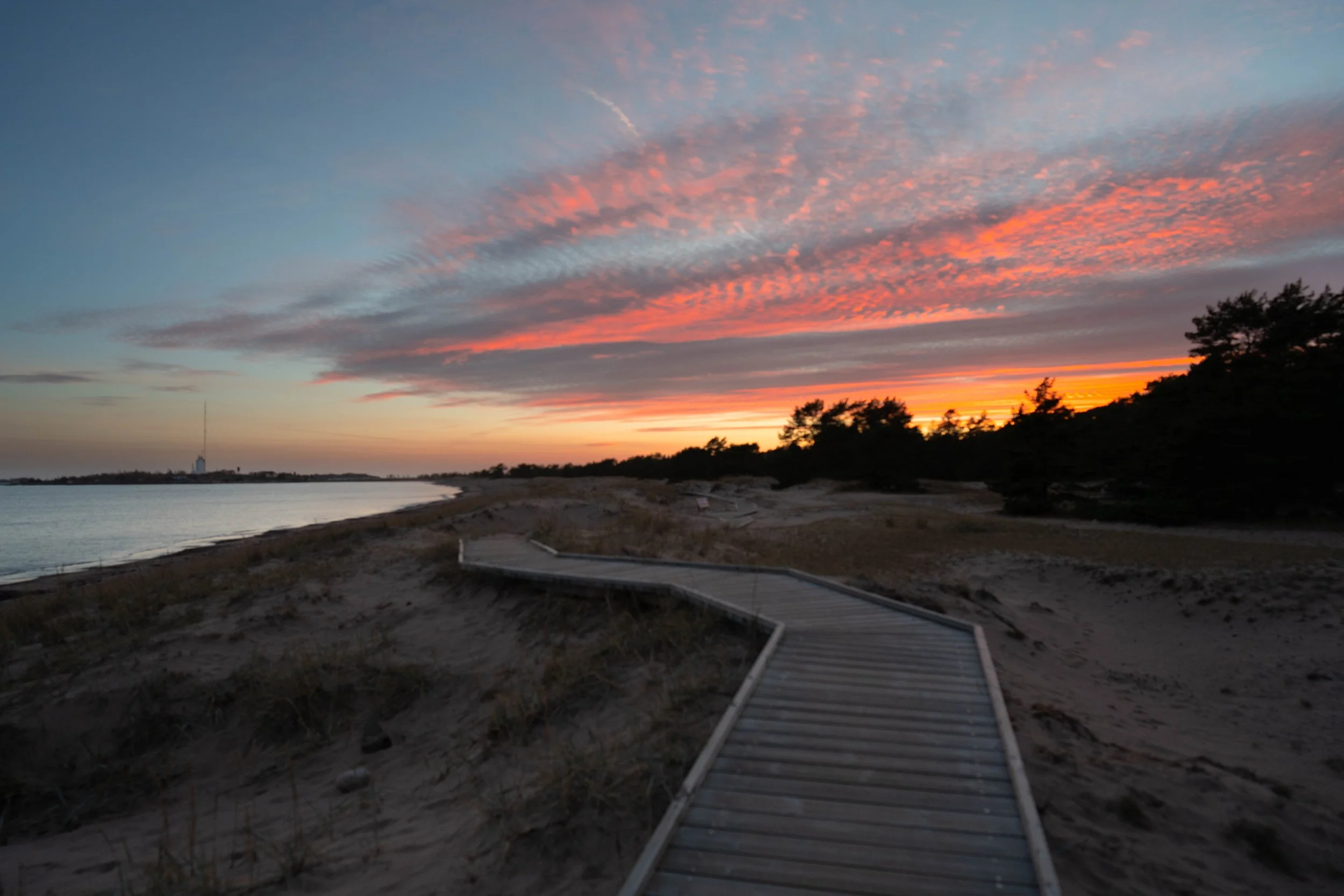 A wooden boardwalk on a sandy beach at sunset, with colorful clouds in the sky and trees in the background.