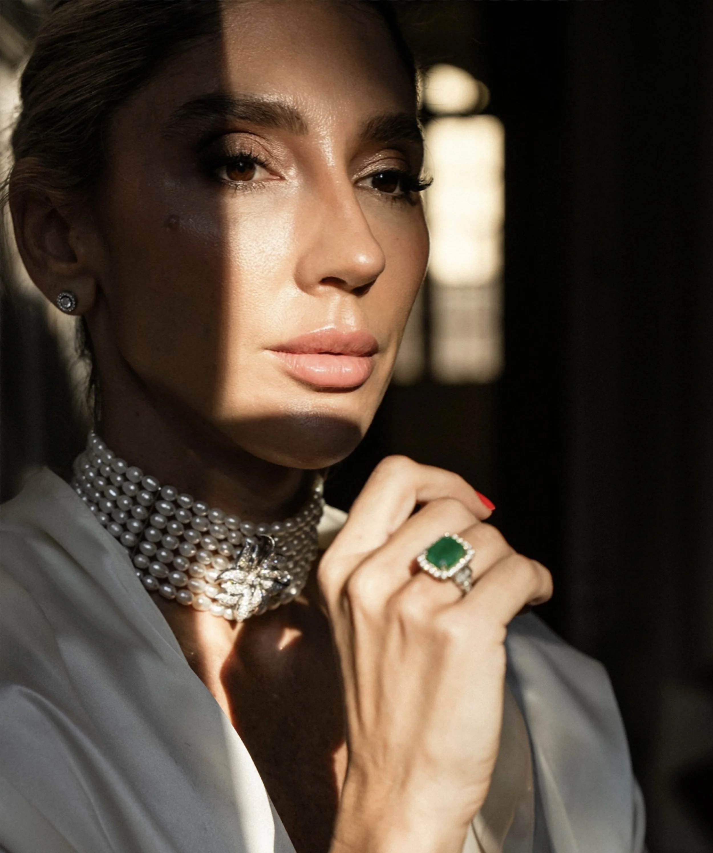 Retrato de mujer con luces y sombras, lleva collar de perlas y anillo con piedra verde, mientras mira pensativa.