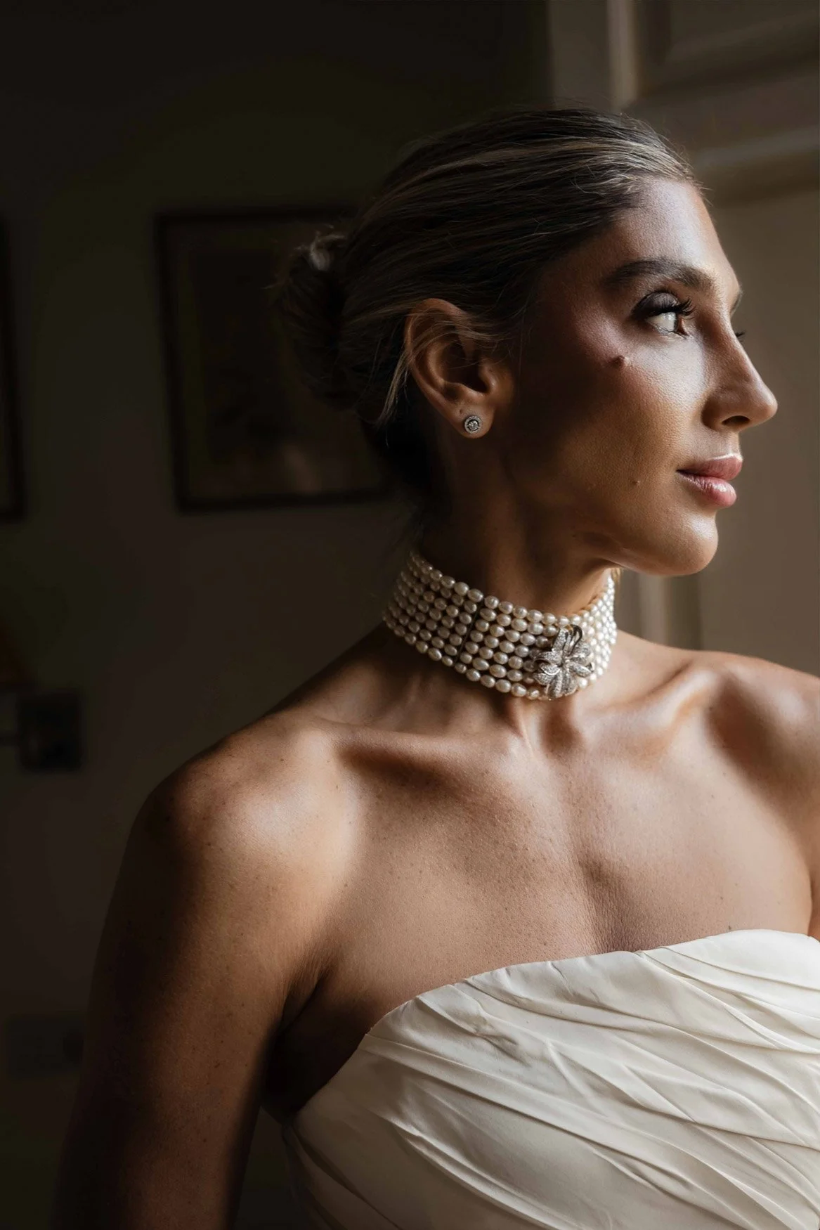 Mujer con vestido de novia y accesorios de perlas, mirando hacia la derecha de la imagen, en un ambiente interior con luz natural.
