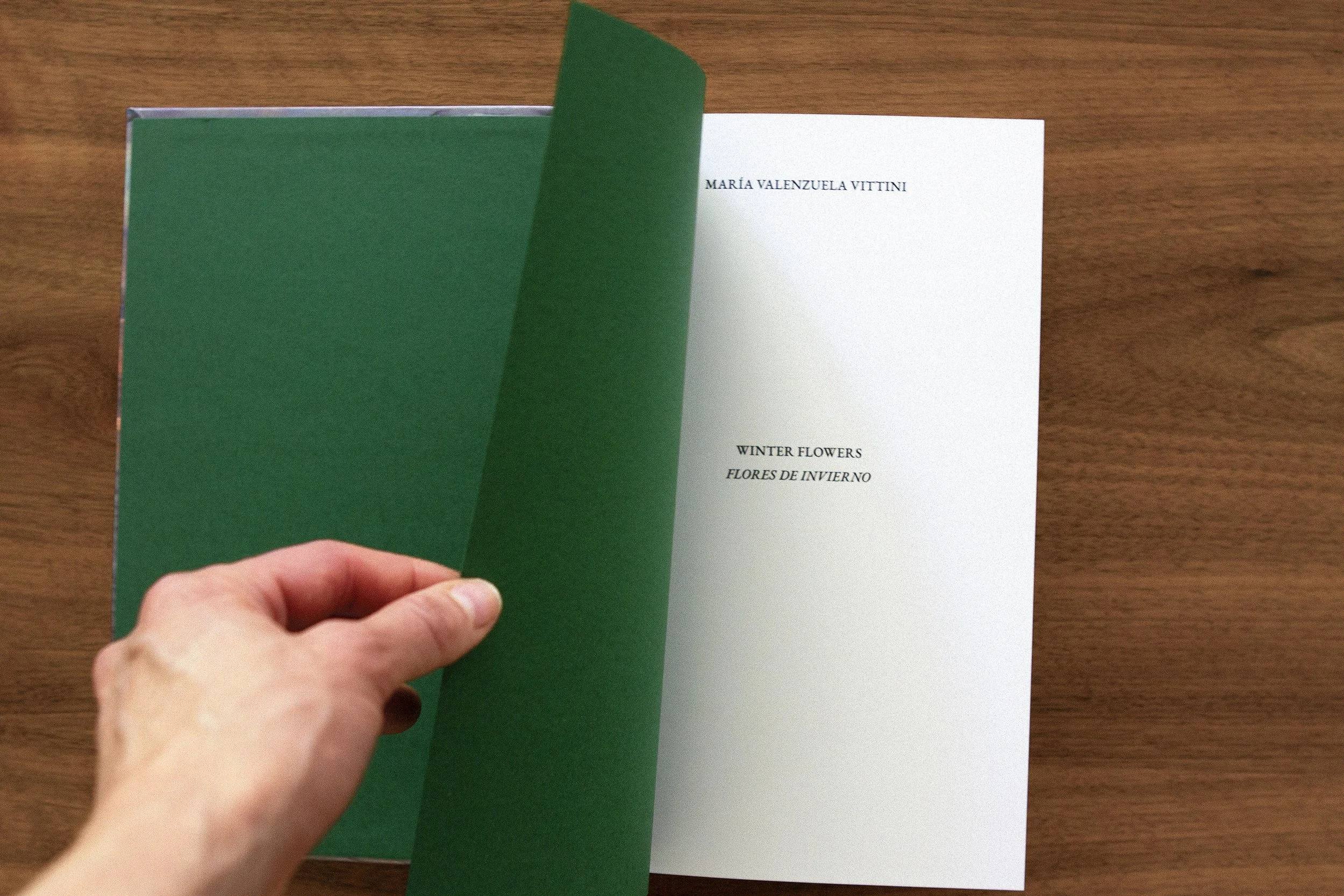 A hand turning the green page of a book on a wooden surface. The open page shows the title ''Winter Flowers'' and ''Flores de Invierno'' with the author's name, María Valenzuela Vittini.