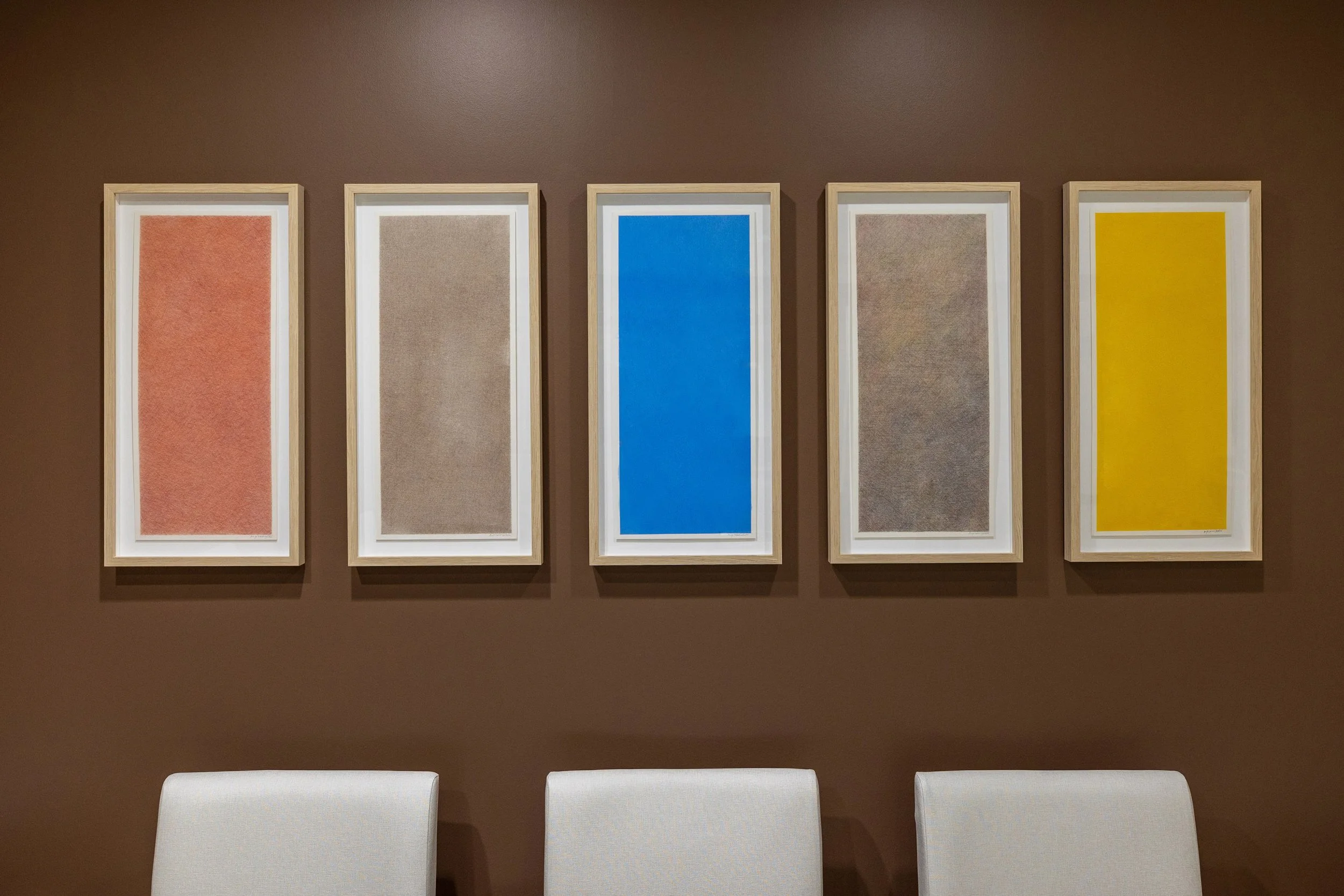 A wall with five framed abstract art pieces in various colors, blue, yellow, and neutral tones.  Color pencil Drawings Commissioned for the collection of The University of Texas SouthWestern. Artist Maria Valenzuela