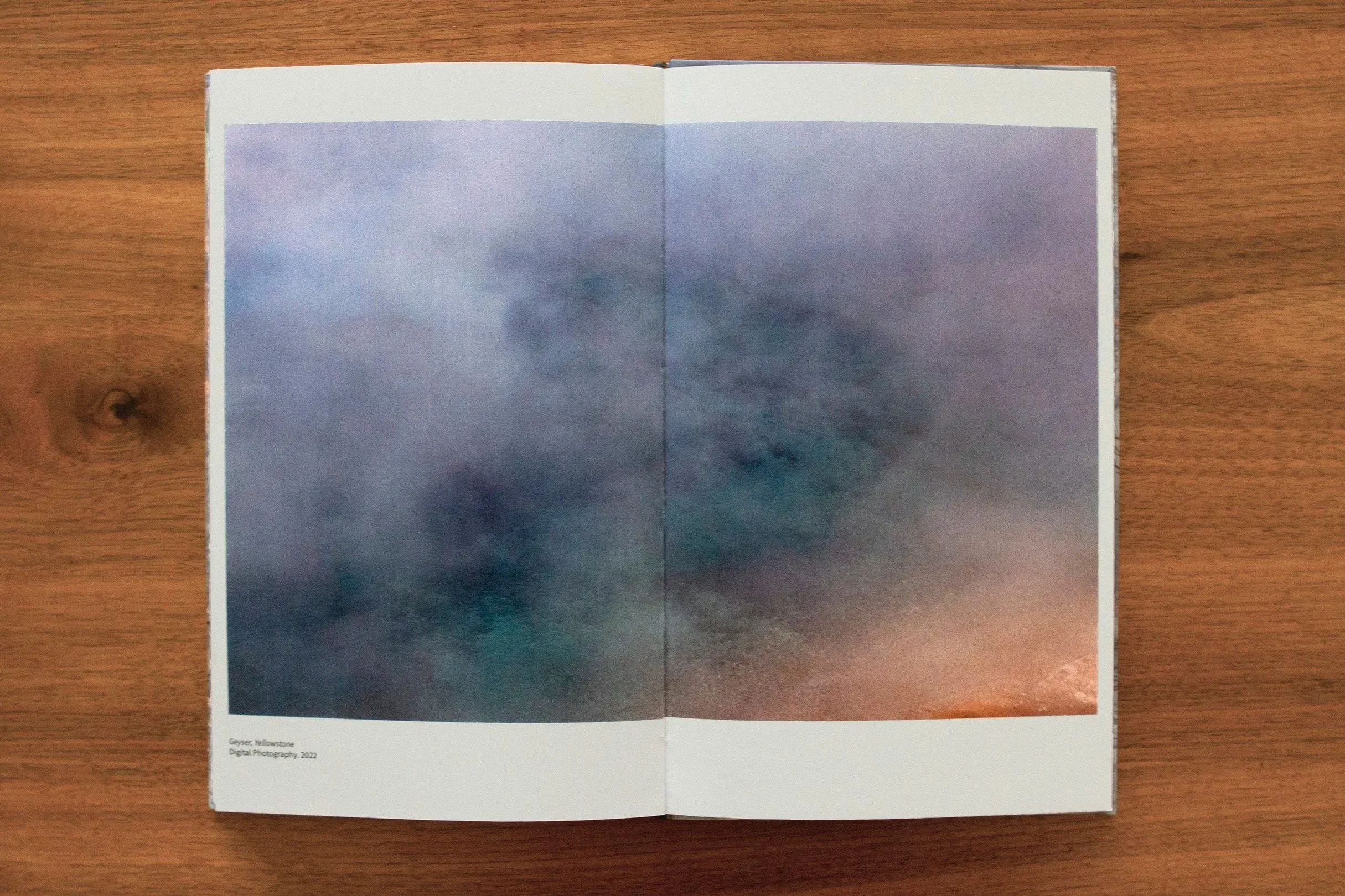 Open booklet on wooden surface shows a sky scene with clouds, labeled 'Geyser, Yellowstone' and attributed to digital photography, 2022.