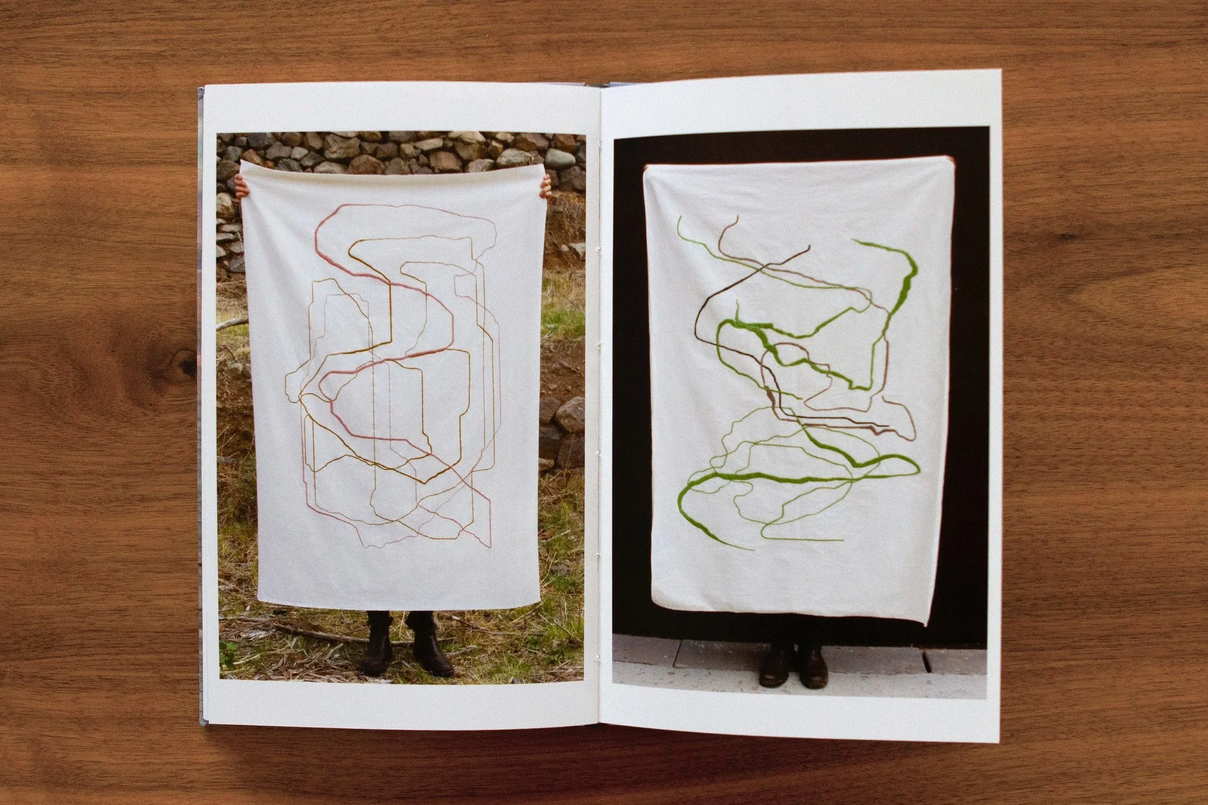 Open book showing two pages with abstract line drawings on fabric stands, outdoors on a wooden surface.