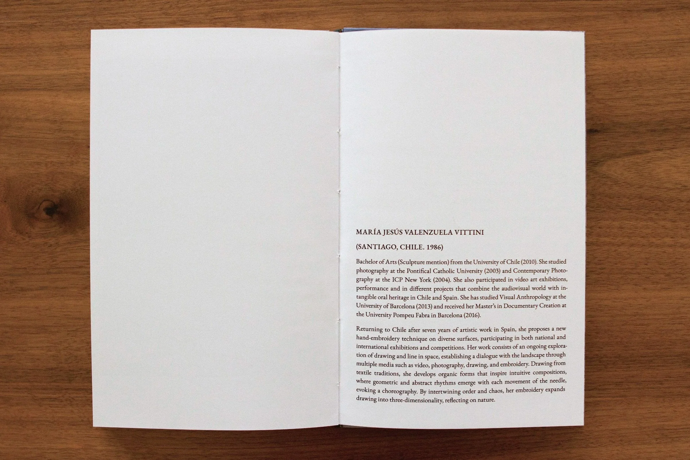 Open booklet placed on a wooden surface, displaying a biography and artwork description of María Jesús Valenzuela Vittini from Santiago, Chile, including her educational background and artistic focus.