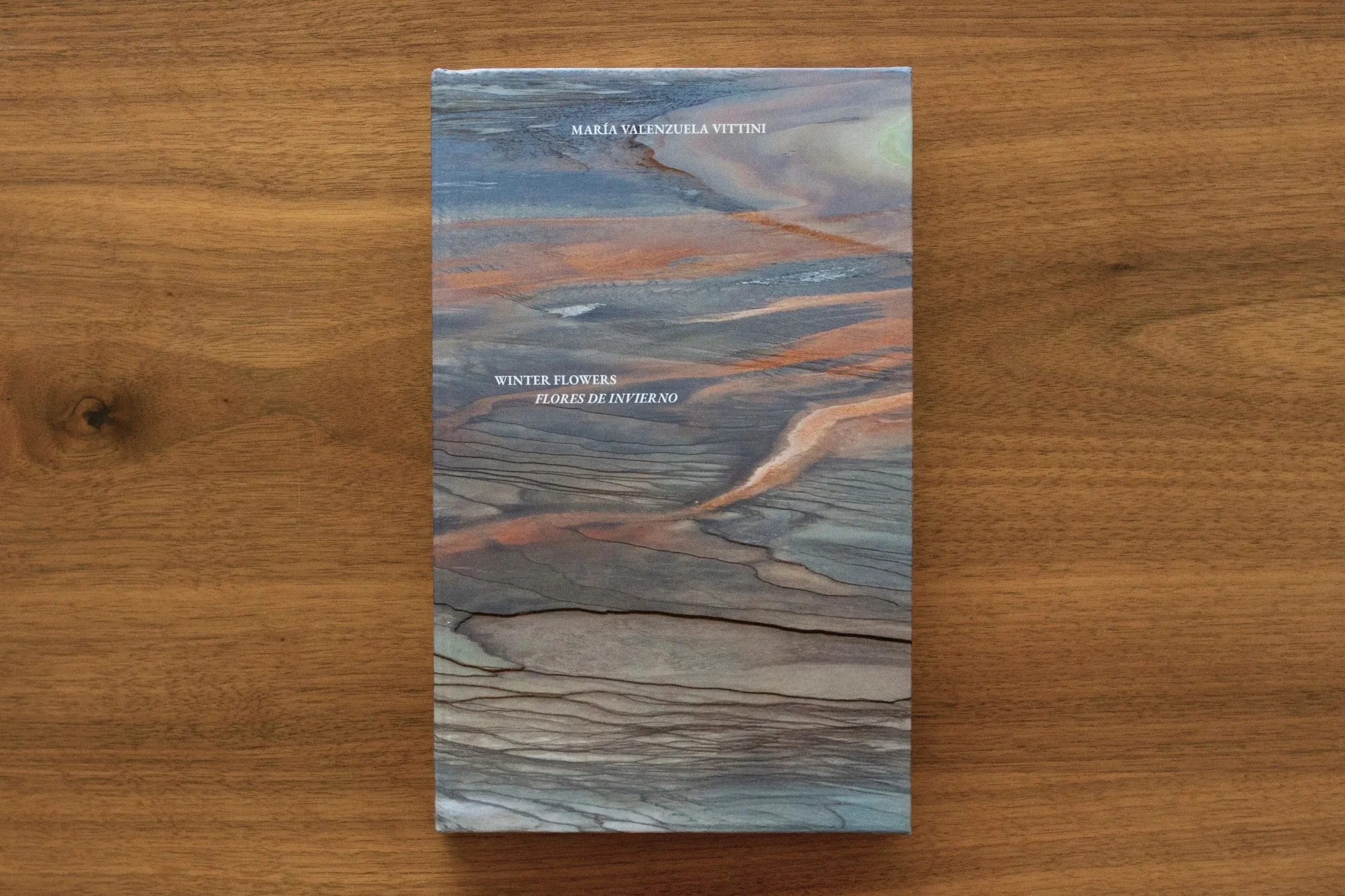 Book titled 'Winter Flowers' by María Valenzuela Vittini on a wooden surface with a colorful, abstract cover design resembling layered mountain landscapes. Catalogo solo show. Libro de artista. Artist Book Maria Valenzuela. Art. Book design.