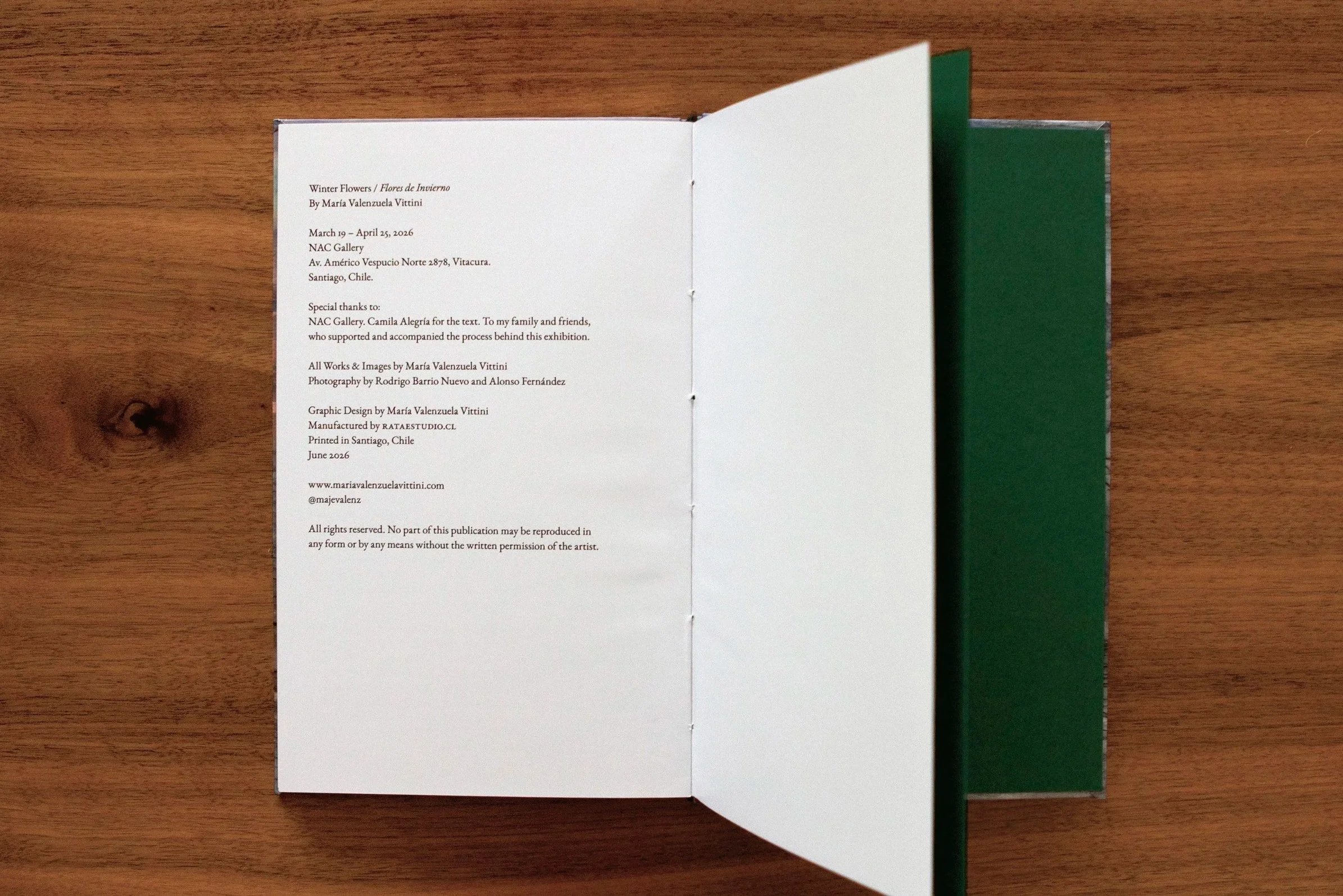 Open book with white pages displaying exhibition information, set on a wooden surface with a green cover visible on the right side.