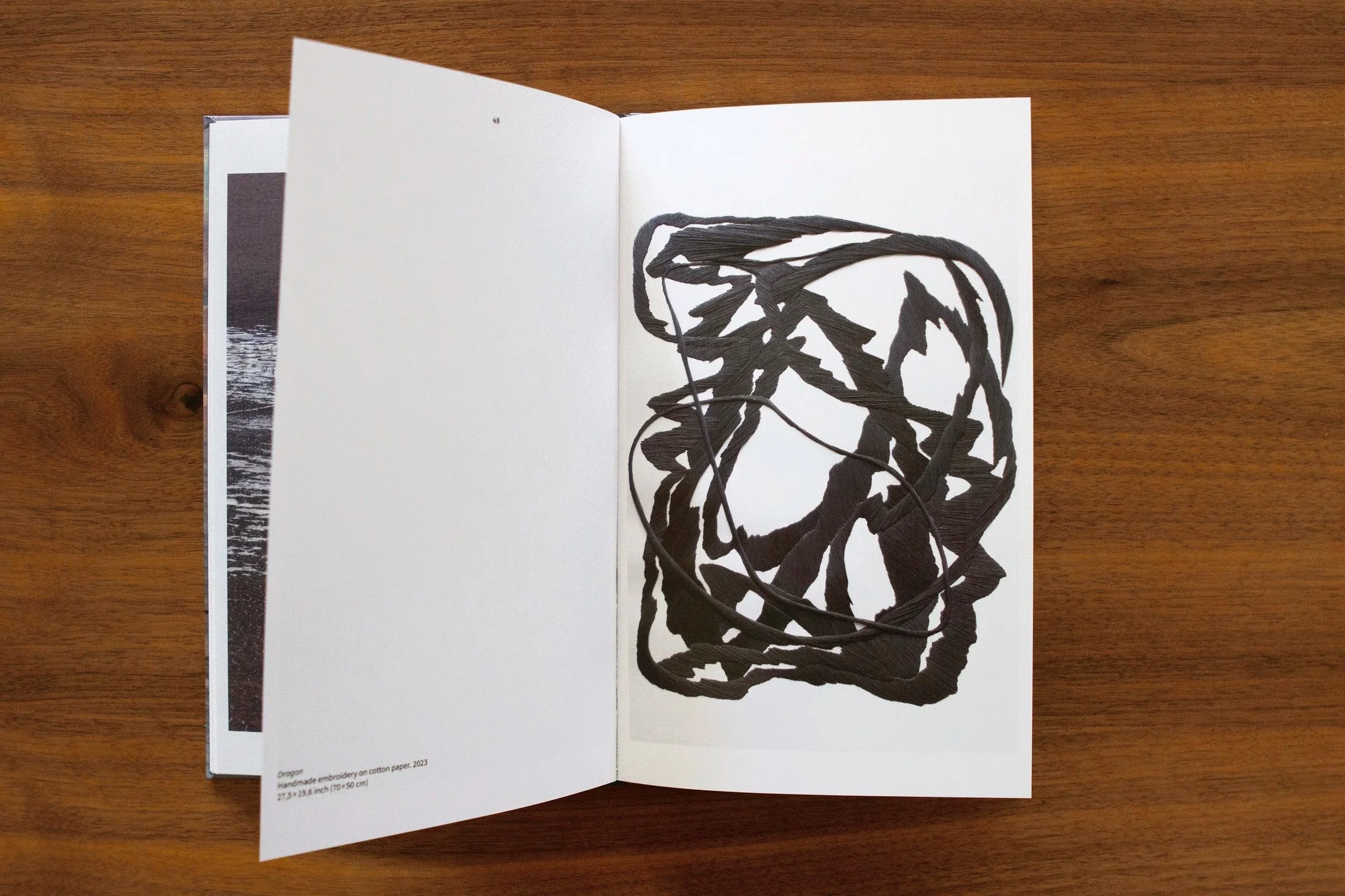 Open art book on a wooden table showing a black ink drawing of a tangled abstract line art. Bordado, hilo, thread, embroidery art. Arte textil Chile.