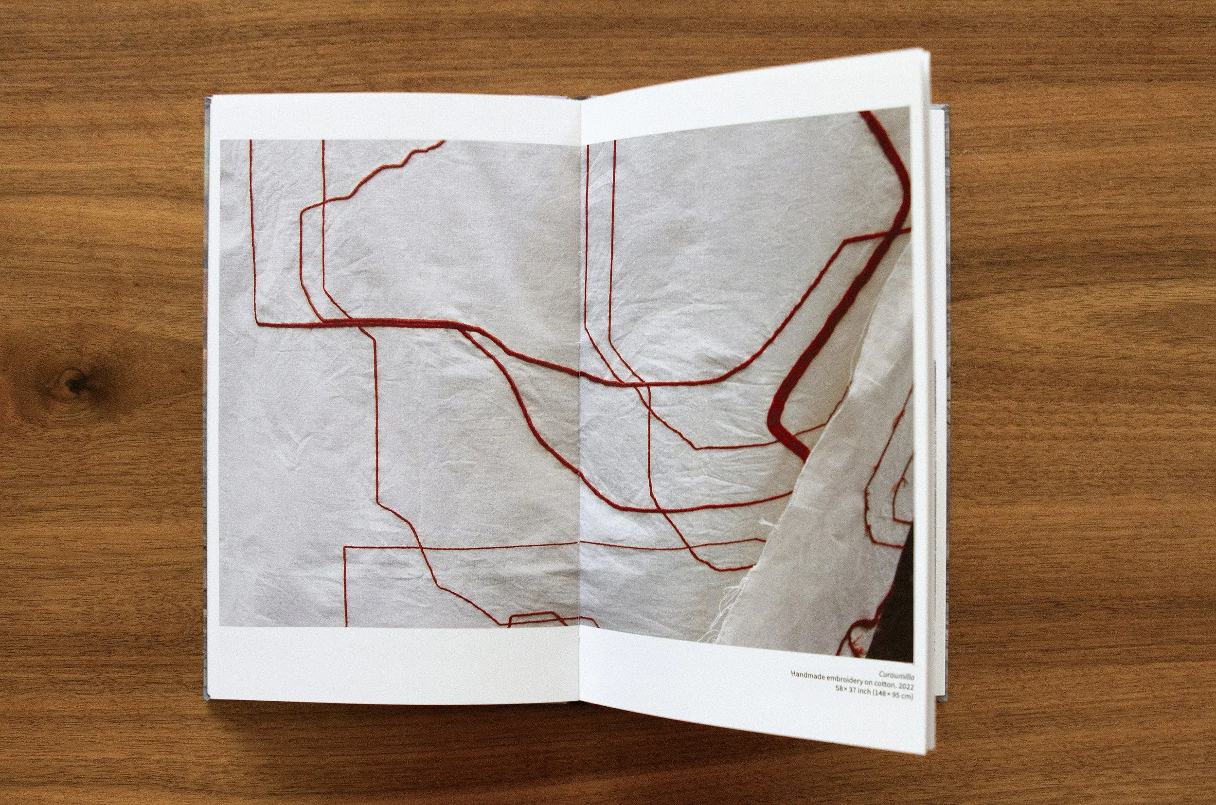 Open book on a wooden surface displaying a fabric with embroidery, featuring red stitched lines. The bottom of the page indicates it is handmade embroidery on cotton, measuring 58 by 37 inches.
