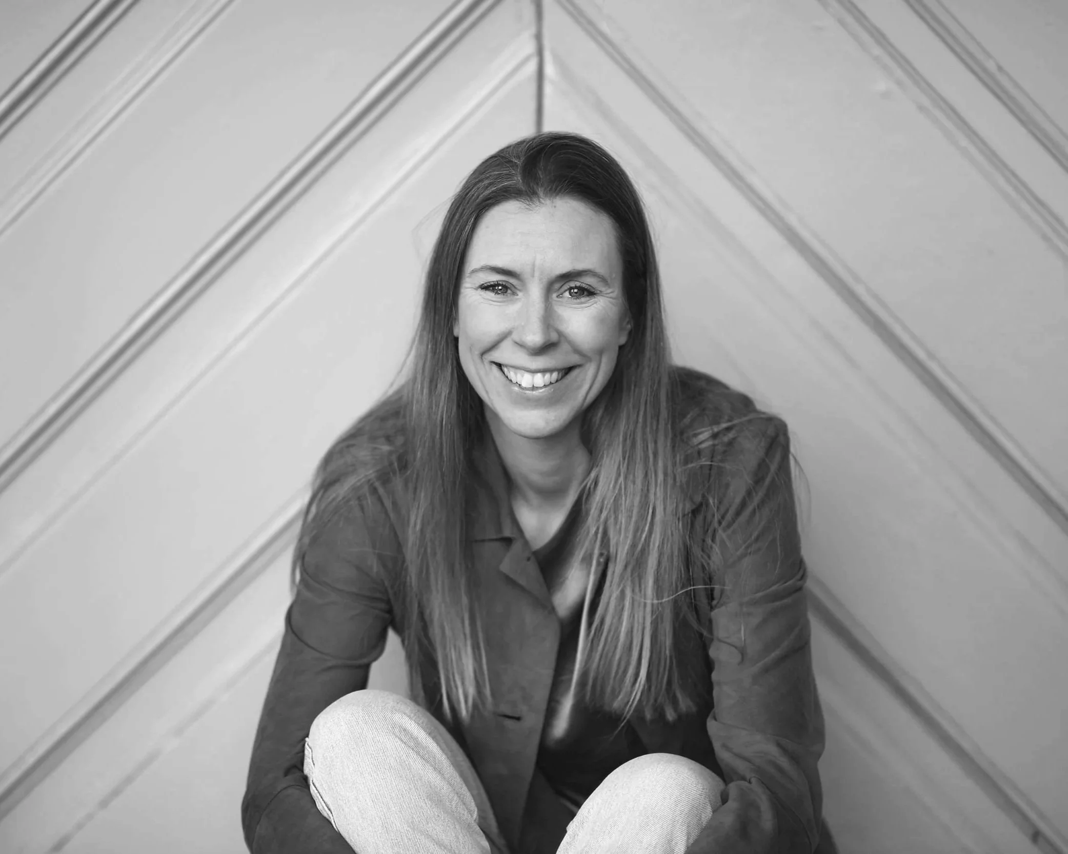 A woman with long hair smiling, sitting on the floor with her knees drawn up, against a background of wooden flooring and diagonal wall paneling.