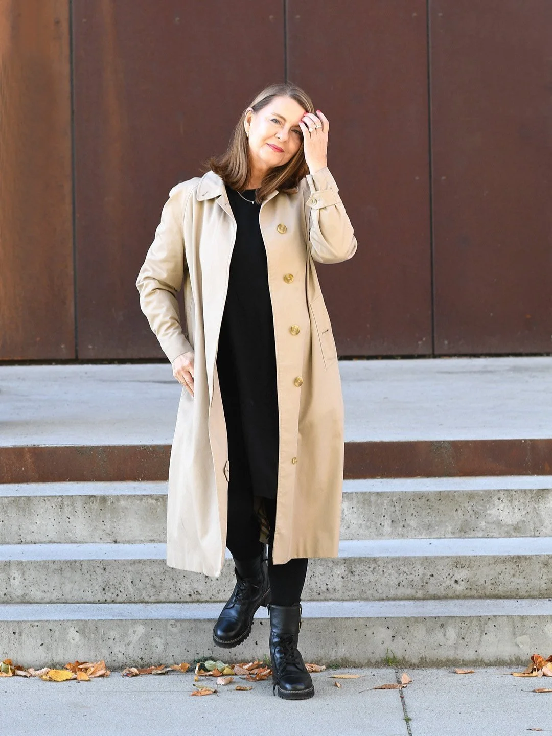 Designcoach Angela Lund i beige frakke på trapper, afslappet stil, sort kjole, støvler, urban baggrund.