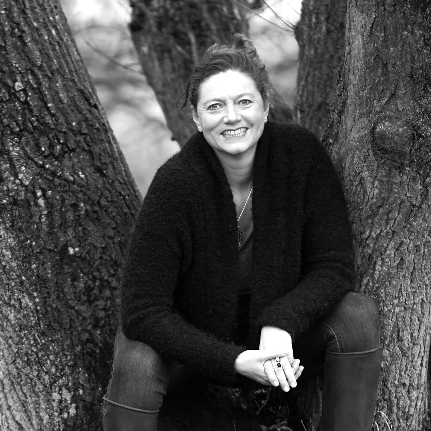 A woman smiling, sitting between two large tree trunks in a natural outdoor setting, in black and white.