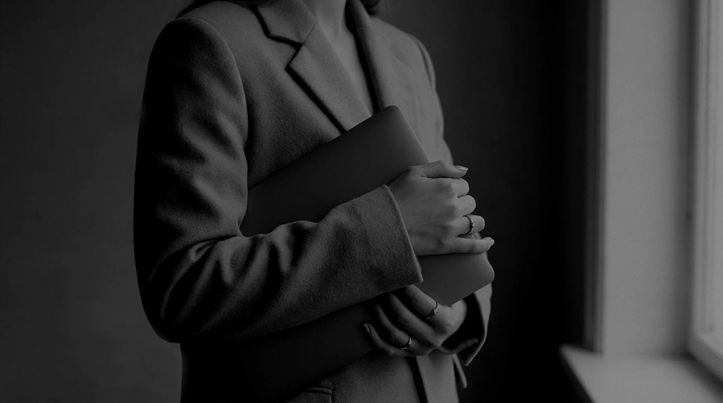 A person in a blazer holding a closed laptop or notebook against their chest, standing near a window with natural light.