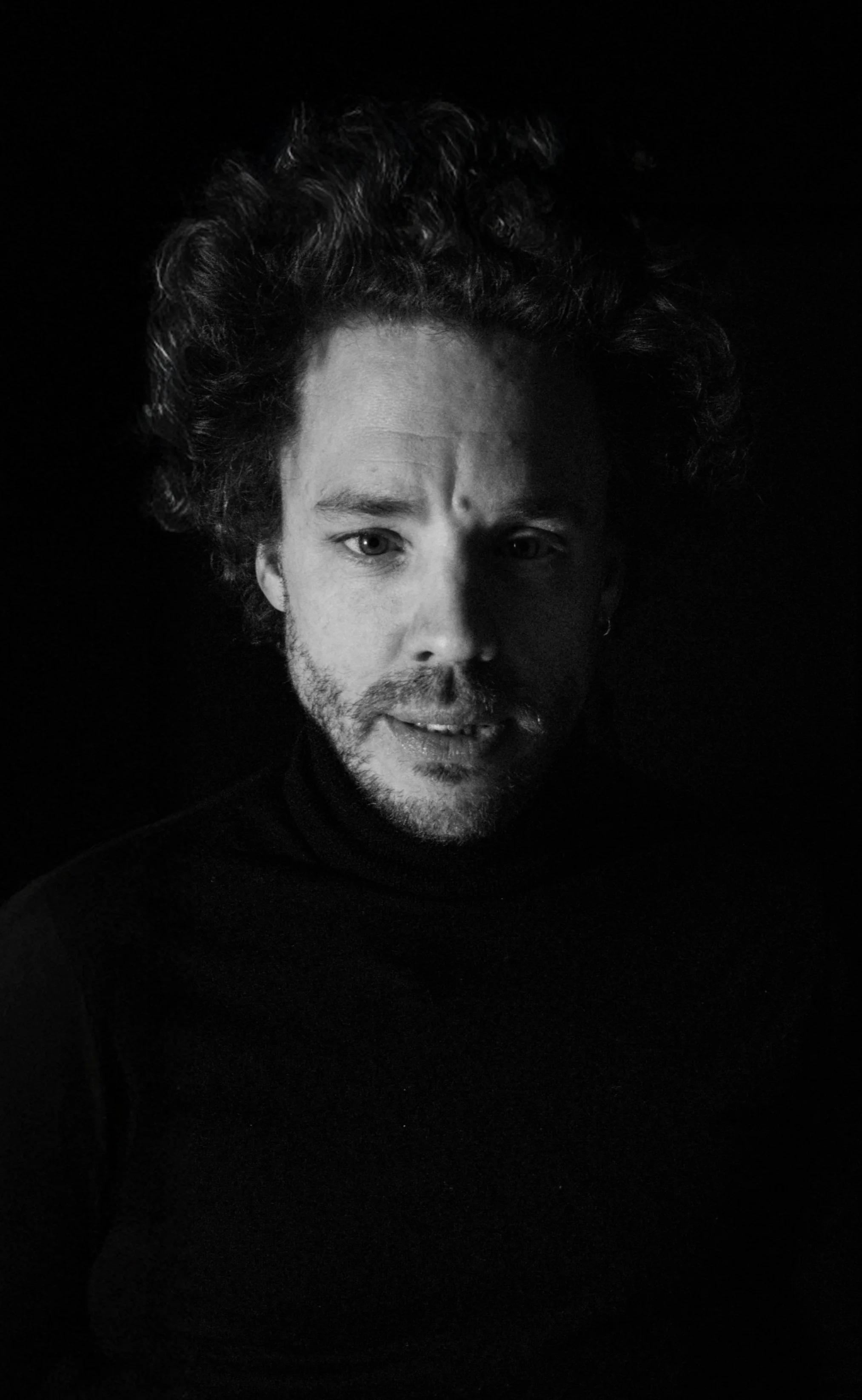 Black and white portrait of a man with curly hair and beard wearing a dark turtleneck